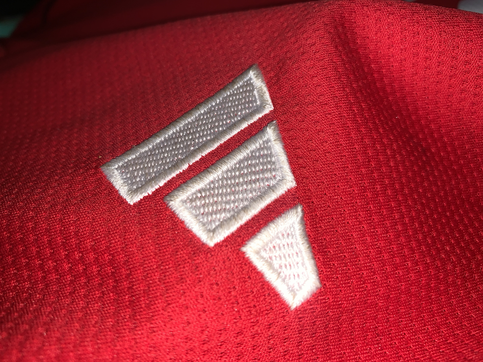 A close-up shot shows the white Adidas logo embroidered on a red fabric. The logo consists of three parallel bars, narrowing from top to bottom. The fabric appears to be textured. No people, specific objects, actions, interactions, or environmental details related to a city or the New Stadium in Nigeria are visible.
