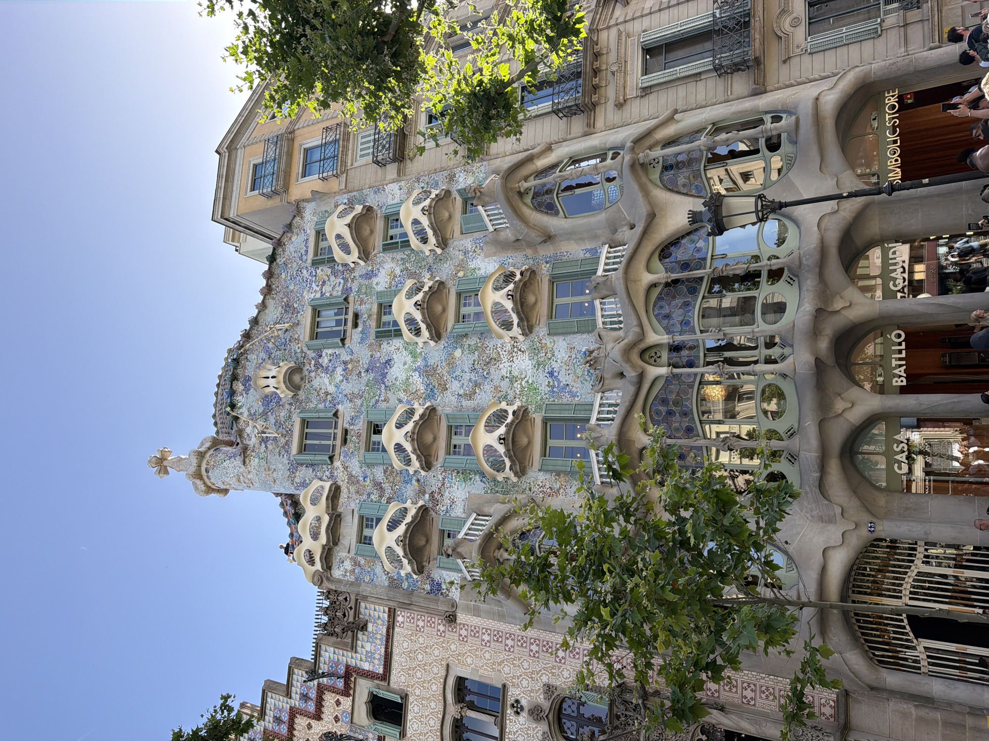 This is a low-angle, outdoor, eye-level shot of Casa Batlló in Barcelona, Spain, on a bright, sunny day. The architecture is the primary subject, with its distinctive facade of colorful mosaic tiles, organic shapes, and bone-like balconies. Lush green trees are visible in the foreground and background, adding a natural element to the urban setting. A clear blue sky fills the upper portion of the frame.
Several people are visible, mostly in the upper right and lower right of the image, appearing to be tourists admiring the building. Some are holding phones and cameras, suggesting they are taking pictures or videos. One person is looking up at the building. The overall mood seems to be one of awe and appreciation for the unique architecture.
Visible text includes "SIMBOLIC STORE" and "CASA BATLLÓ" on signage near the entrance of the building. The number "43" is also visible on a small plaque near the entrance. The architecture is characteristic of Antoni Gaudí's work, known for its surreal and organic forms. The time of day is likely mid-morning or afternoon, given the bright sunlight and clear sky.