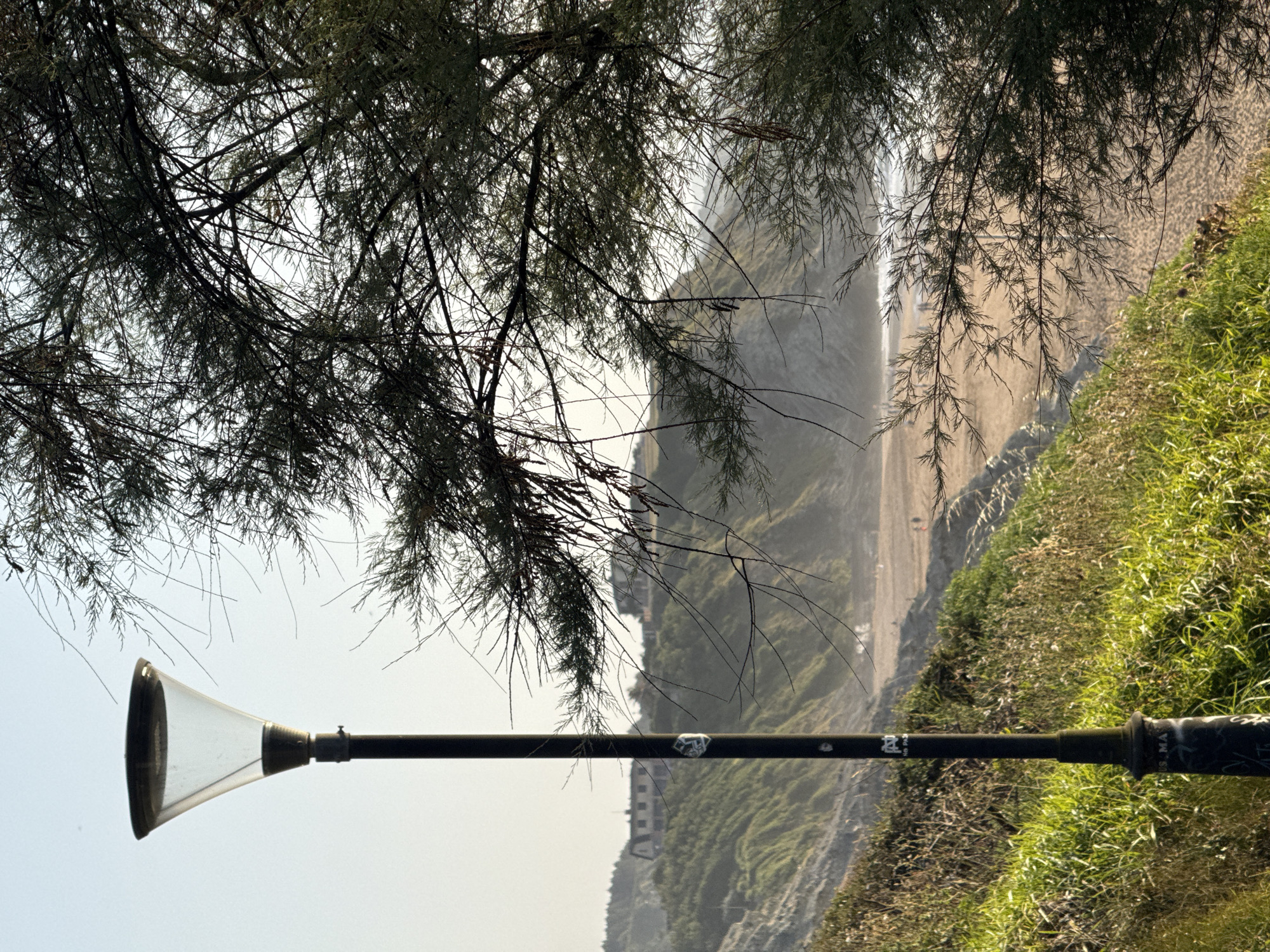 The image depicts a view of the Barinatxe beach in Sopela, Spain, seen from an elevated grassy area. In the foreground, a dark lamppost stands vertically on the left, with a clear glass top and a black pole. Graffiti is visible on the base of the lamppost. To the right, the branches of a dark green tree with thin needles partially obscure the view of the beach and the sea.
The midground shows a sandy beach with a few distant figures walking. The coastline is characterized by rolling green hills and cliffs. In the background, several buildings are visible atop the hills. The sky is a pale, hazy blue, suggesting either early morning or late afternoon light. The overall atmosphere is serene and natural.