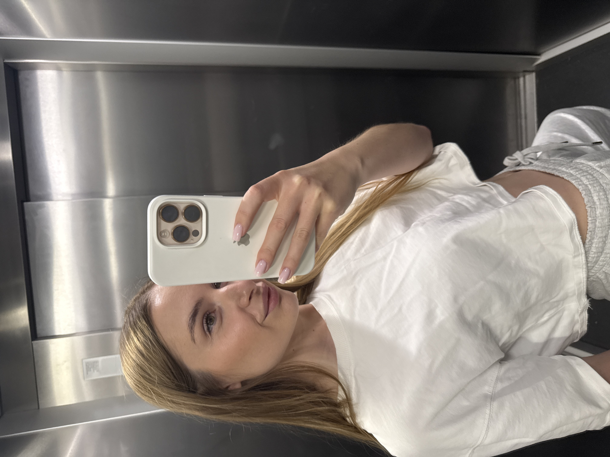 A woman is taking a selfie with a white iPhone in an elevator. The elevator has stainless steel walls. The woman has blonde hair and is wearing a white t-shirt and gray shorts. She is facing the camera and puckering her lips. The location is likely in Budva, Montenegro, but this is not visible in the image.