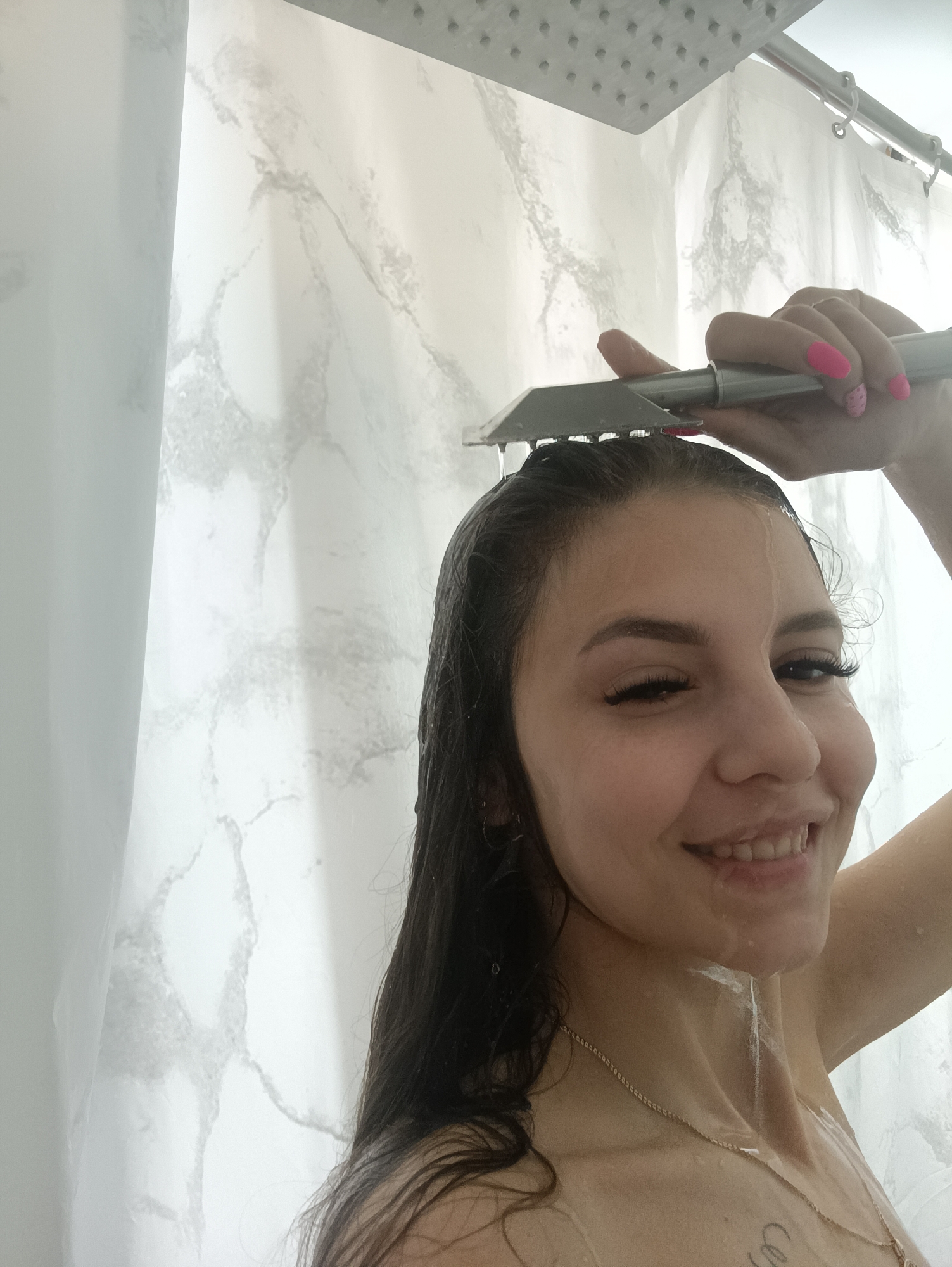 A woman with wet, dark hair is in a shower. She is holding a handheld shower head over her head, and water is running down her face and body. She is smiling and looking at the camera. She is wearing a gold necklace. The shower has a marble-patterned curtain. The location is Unknown, Unknown.
