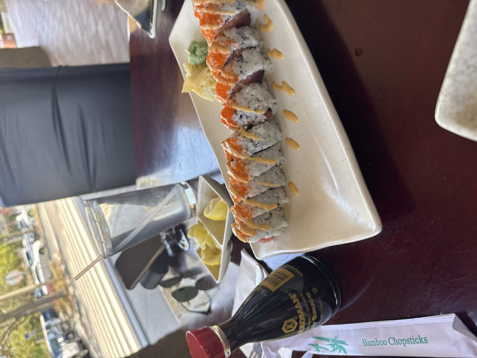 A high-angle interior photograph depicts a sushi meal served on a dark brown wooden table within a restaurant in Naples, United States. The central focus is a long, off-white rectangular plate holding ten pieces of sushi. Each sushi piece contains white rice, a dark red interior (consistent with tuna), is topped with orange roe, and is drizzled with a light orange sauce. The plate also features a small mound of pickled ginger and a dollop of green wasabi. To the left, a dark brown Kikkoman soy sauce bottle with a red cap stands beside a white paper sleeve labeled "Bamboo Chopsticks." Further left, a white dish with two compartments holds lemon wedges. Behind these items, a clear glass of water containing a white straw, a dark-colored smartphone, and dark sunglasses are present on the table. In the blurred background, a dark gray chair back is visible, with a large window revealing a sunlit exterior street scene featuring a sidewalk, parked vehicles, a road, and trees. No individuals are present in the frame.