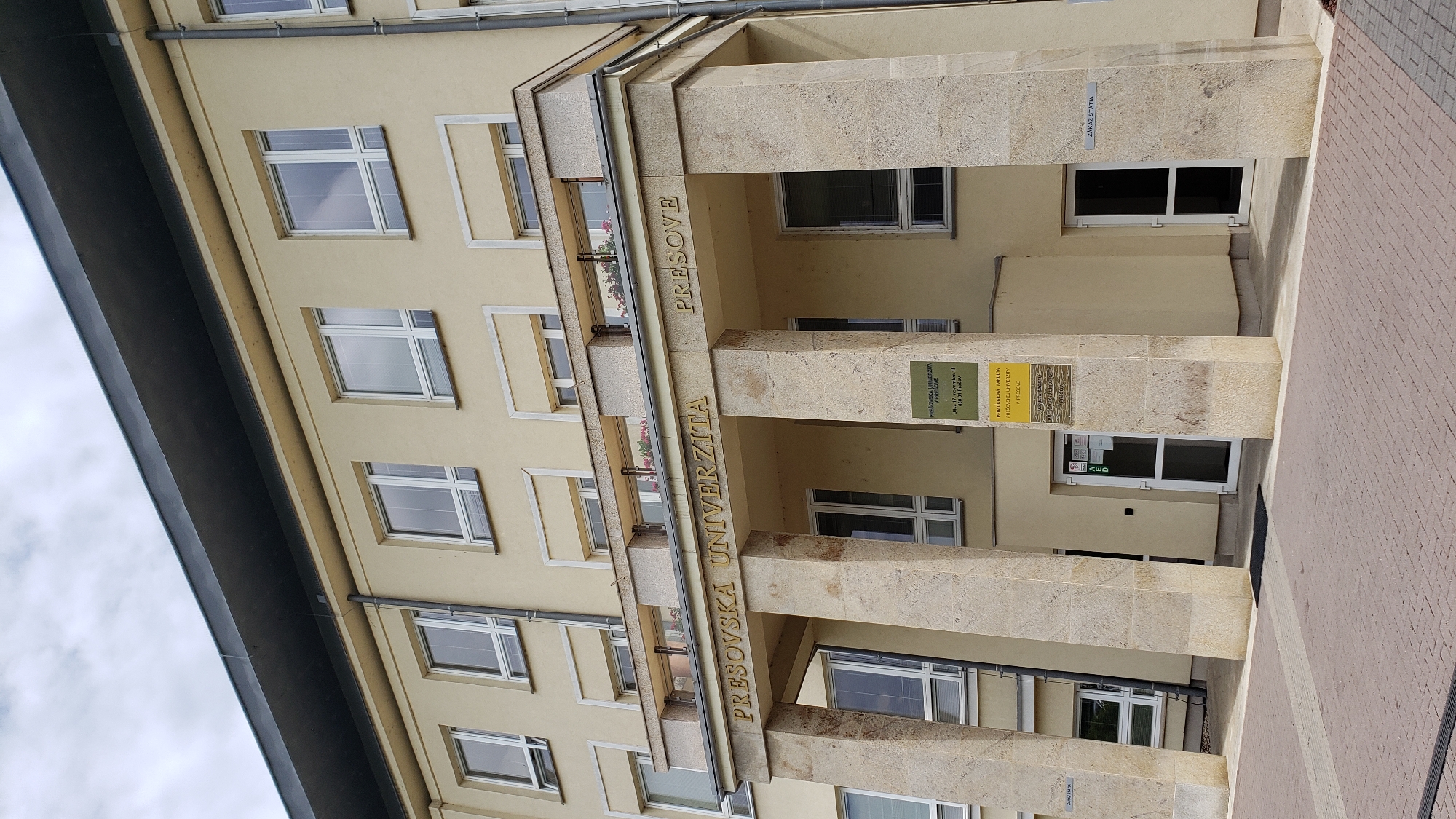 The image captures a head-on view of the main facade of the University of Prešov's Faculty of Education building in Prešov, Slovakia, located at Ul. 17. novembra 15, 080 01 Prešov. The multi-story building features a light yellow-cream stucco exterior under an overcast sky.
The prominent entrance consists of a portico supported by four large, light-colored stone columns. Above these columns, golden three-dimensional letters are affixed to the stone structure, spelling "PREŠOVSKÁ UNIVERZITA" on the left and "PREŠOVE" on the right. Above this lettering, a balcony or ledge spans the width of the portico, featuring decorative railings and planters containing red flowers.
The building's upper floors display numerous rectangular windows with white frames, many of which contain horizontal blinds. These windows are evenly spaced across the facade. A dark, flat roof overhang is visible at the top of the building. A grey downspout runs vertically along the right side of the building's exterior.
At ground level, below the portico, are two white-framed glass doors. The door on the left has a red "AED" sign affixed to it. To the right of the rightmost column, a small white sign with black text reads "ZÁKAZ STÁTIA" (No Stopping/Parking). A partially visible sign to the left of the leftmost column also appears to read "ZÁKAZ STÁTIA".
Multiple informational signs are mounted on the wall to the right of the left entrance door. The top green sign confirms "PREŠOVSKÁ UNIVERZITA V PREŠOVE" and provides the address "Ul. 17. novembra 15, 080 01 Prešov". Below it, a yellow sign identifies the "PEDAGOGICKÁ FAKULTA PREŠOVSKEJ UNIVERZITY V PREŠOVE" (Faculty of Education of the University of Prešov in Prešov). A third, darker sign below the yellow one indicates the "ÚSTAV TELESNEJ VÝCHOVY A ŠPORTU" (Institute of Physical Education and Sport).
The foreground features a paved area constructed with interlocked red and grey pavers. A black doormat is placed directly in front of the left entrance door. No individuals or vehicles are present in the scene.