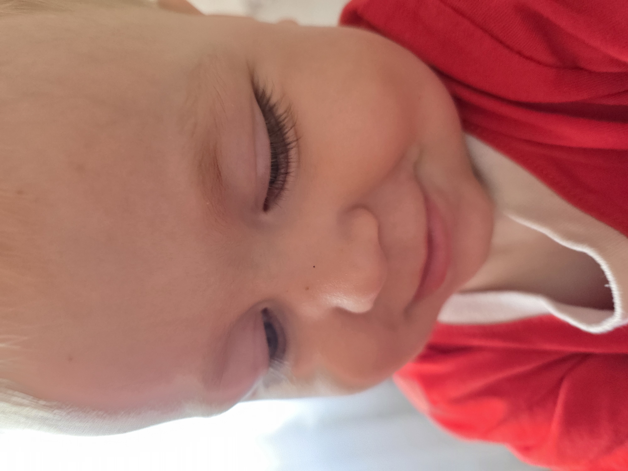 A close-up, slightly angled view of a baby with fair skin, lying down. The baby's eyes are closed and long eyelashes are visible. The baby has a small mole on its nose. The baby is wearing a red shirt with a white trim. The location is not specified.