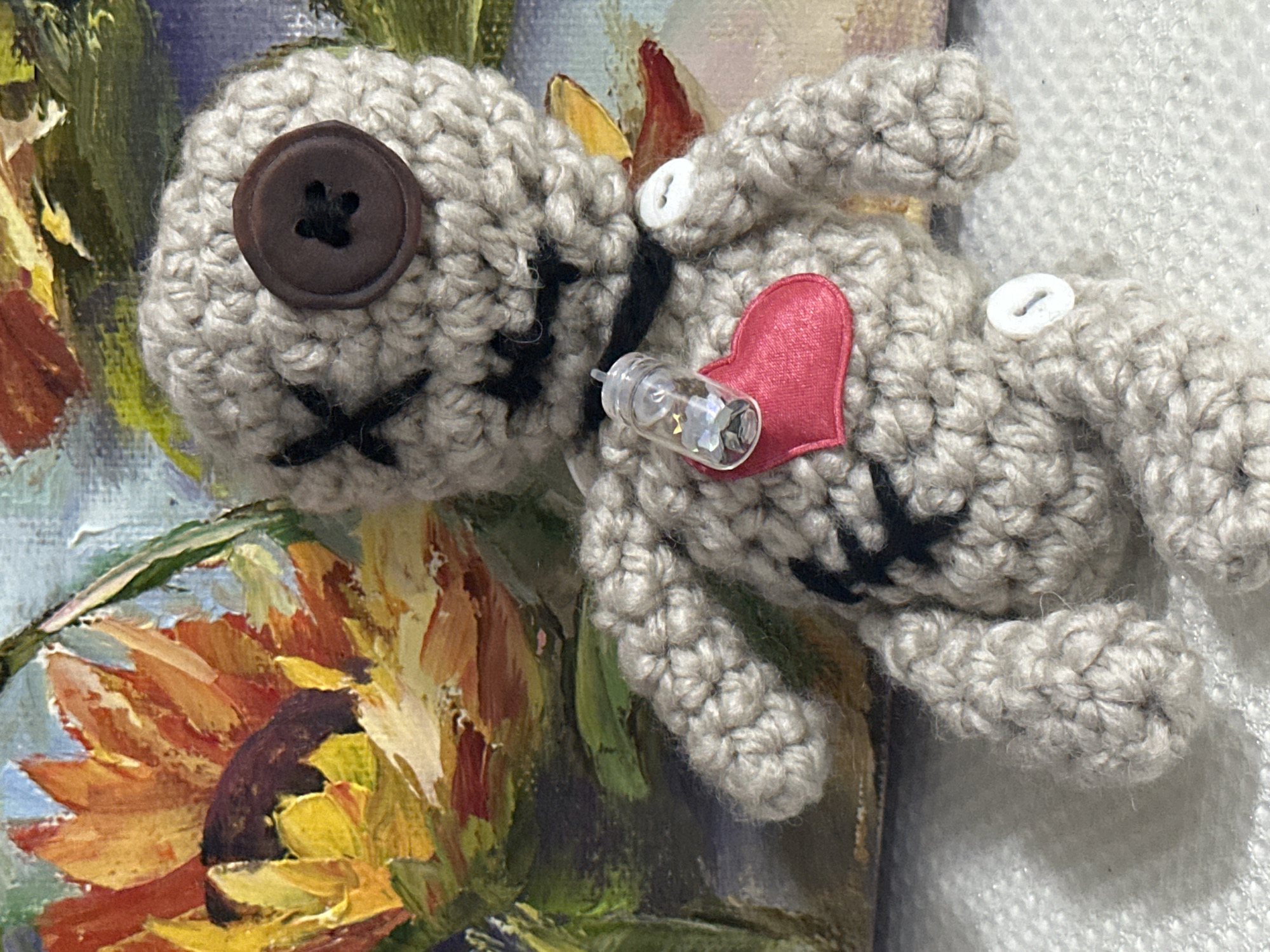 This is a close-up, eye-level, indoor photograph of a handmade crochet doll. The doll is a light gray color and has a button for one eye, with stitched black Xs for the other eye and mouth. It has a small glass vial containing glittering fragments around its neck, and a red felt heart sewn onto its chest. A white button is visible on its right arm, and another on its lower body. The background is a painting of a sunflower. The image does not contain any information about the city of Nevinnomyssk, Russia, or any other specific location.