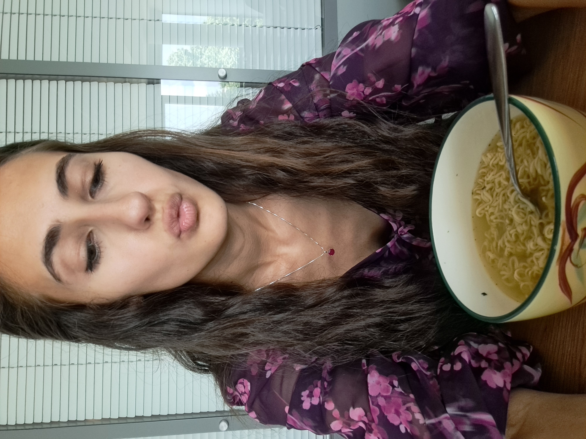 A young woman with long, dark, wavy hair is captured in a playful indoor portrait. She is wearing a sheer, dark purple top adorned with a pink floral pattern and a delicate silver necklace featuring a small red pendant. Her eyes are partially closed, and her lips are pursed in a "duck face" expression, directed towards the camera.
Beside her, on a light wooden surface, sits a yellow and cream-colored bowl with a green rim and reddish-brown abstract designs. The bowl contains instant ramen or noodles in a clear broth, with a metal spoon resting inside.
The background features white horizontal window blinds, through which diffused daylight and faint glimpses of outdoor greenery are visible, suggesting a daytime setting in an urban environment like Sofia, Bulgaria. The overall impression is candid and casual. No visible text is present in the image.