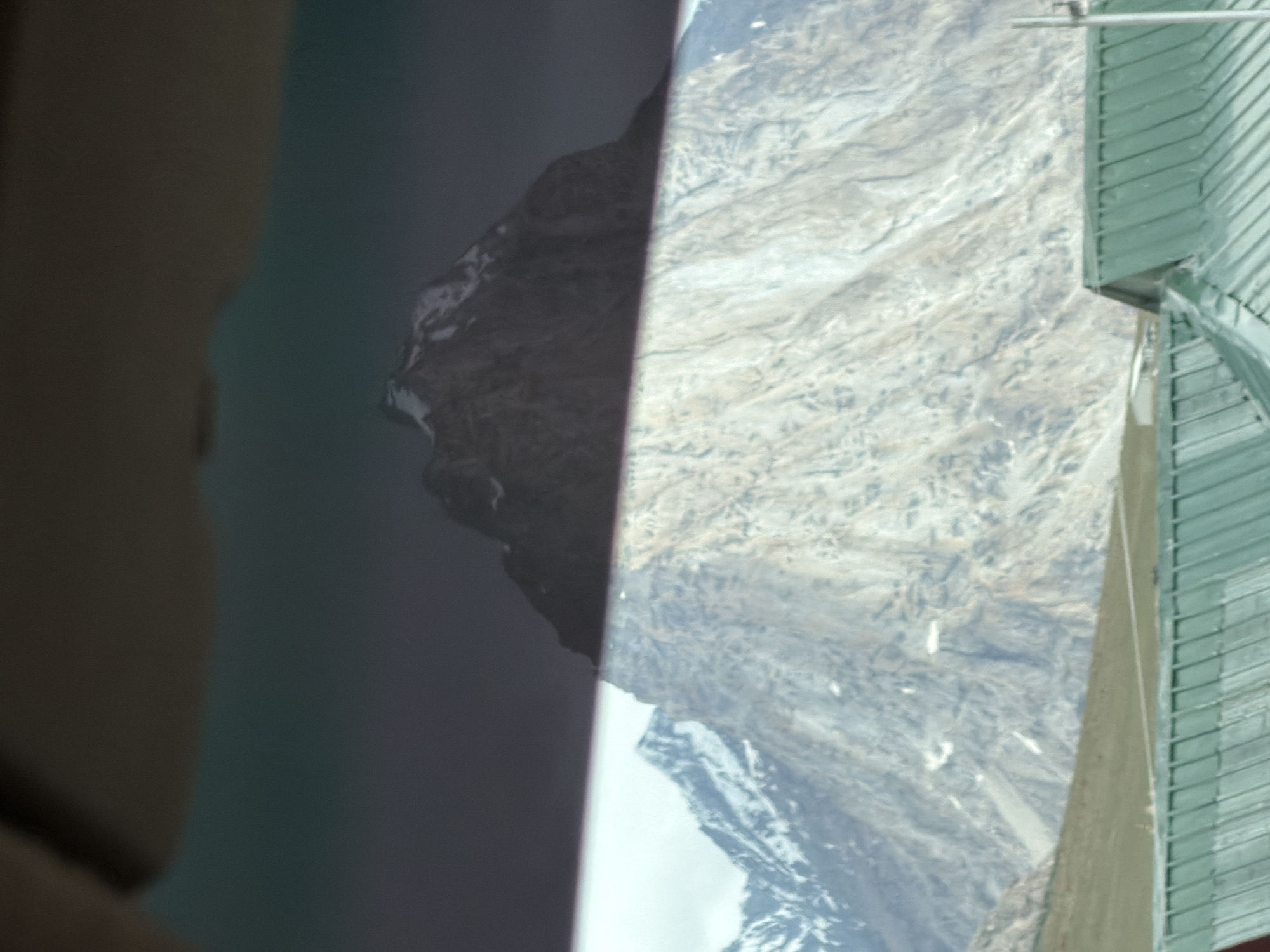A mountainous landscape is depicted, with snow-capped peaks in the distance. In the foreground, there is a green, corrugated metal roof of a building. The image appears to be taken from inside a vehicle, with a dark interior element visible at the top. The location is Kurgiakh, India.