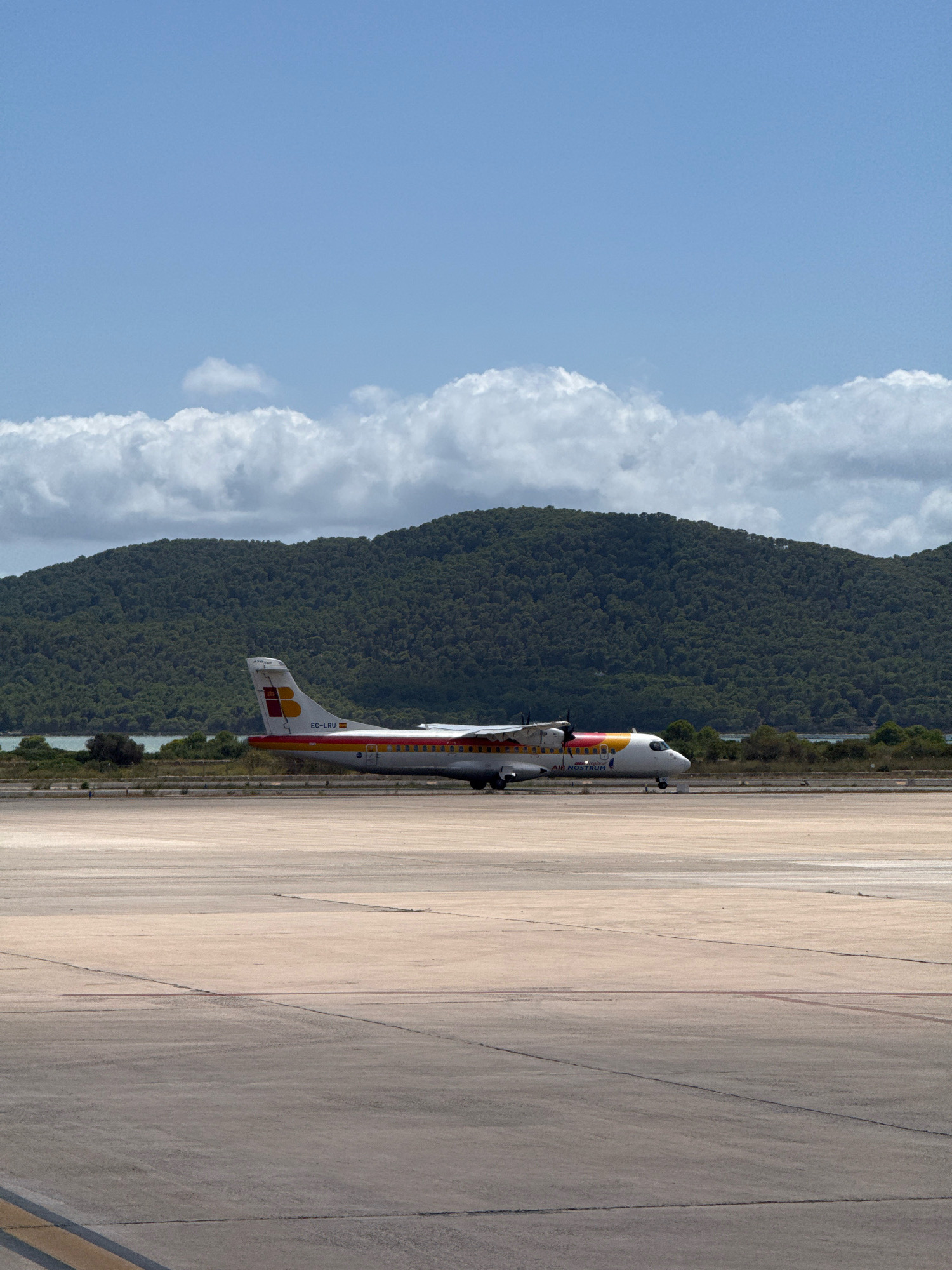 The image depicts an Air Nostrum turboprop airplane parked on a tarmac at Ibiza Airport (IBZ), located in Sant Josep de sa Talaia, Spain. The airplane, with registration EC-LRU, is positioned on a light-colored concrete surface, with tire marks visible on the ground. In the background, a body of water is situated behind a line of trees and shrubs. Further in the distance, a verdant, tree-covered hill rises, and a bright blue sky with scattered white clouds forms the upper portion of the frame. The scene suggests a clear day, likely at an airport in a coastal or island region.