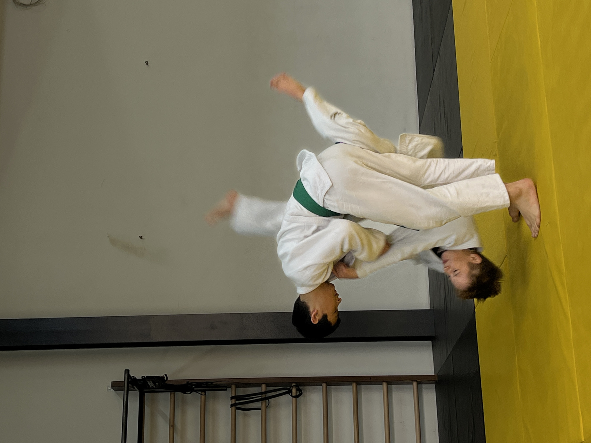 Two people are practicing judo on a mat. One person, wearing a white judogi with a green belt, is throwing the other person, who is also wearing a white judogi. The scene is indoors, with walls, a wooden rack, and mats visible. The mat colors are black, yellow, and gray. The location is not specified, and therefore cannot be attributed to Krasnoyarsk, Russia.
