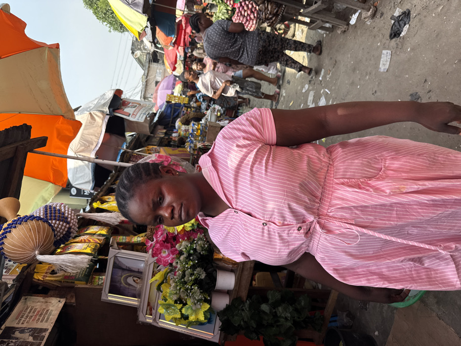 The image captures a vibrant outdoor market scene at Iya Adun Market on Sura Bale Street, located within the Lekki area of Lagos, Nigeria.
In the foreground, a dark-skinned woman with tightly braided hair stands, her gaze directed slightly to the left. She wears a short-sleeved, collared dress of pink fabric with thin white vertical stripes. The dress features buttons down the front, a drawstring at the waist, and two visible pockets. Linear scars or marks are visible on her right forearm. A light yellow stain is present on the lower right side of her dress.
To the immediate left and behind the woman, a market stall is visible. It displays artificial flowers in shades of yellow and pink, arranged in white pots, alongside several framed pictures, one of which clearly depicts a religious image resembling the Virgin Mary. Above these, traditional beaded percussion instruments, similar to shekeres, and yellow packaged goods hang. A black and white sign affixed to this stall reads: "SURA BALE STREET, IYA ADUN MARKET, EPE-OSA, LAGOS."
The background shows a crowded market alleyway under a clear, bright sky. Multiple individuals, predominantly women, are observed walking and standing among numerous market stalls. These stalls are shaded by a mosaic of large umbrellas in various colors, including orange, white, yellow, pink, and blue, interspersed with corrugated metal roofing. Produce, specifically mounds of red items resembling onions, is displayed on wooden stands. The ground surface is a mixture of dirt and worn concrete, marked by scattered litter such as plastic bags and wrappers. Distant buildings and overhead electrical wires are visible beneath the bright daylight.