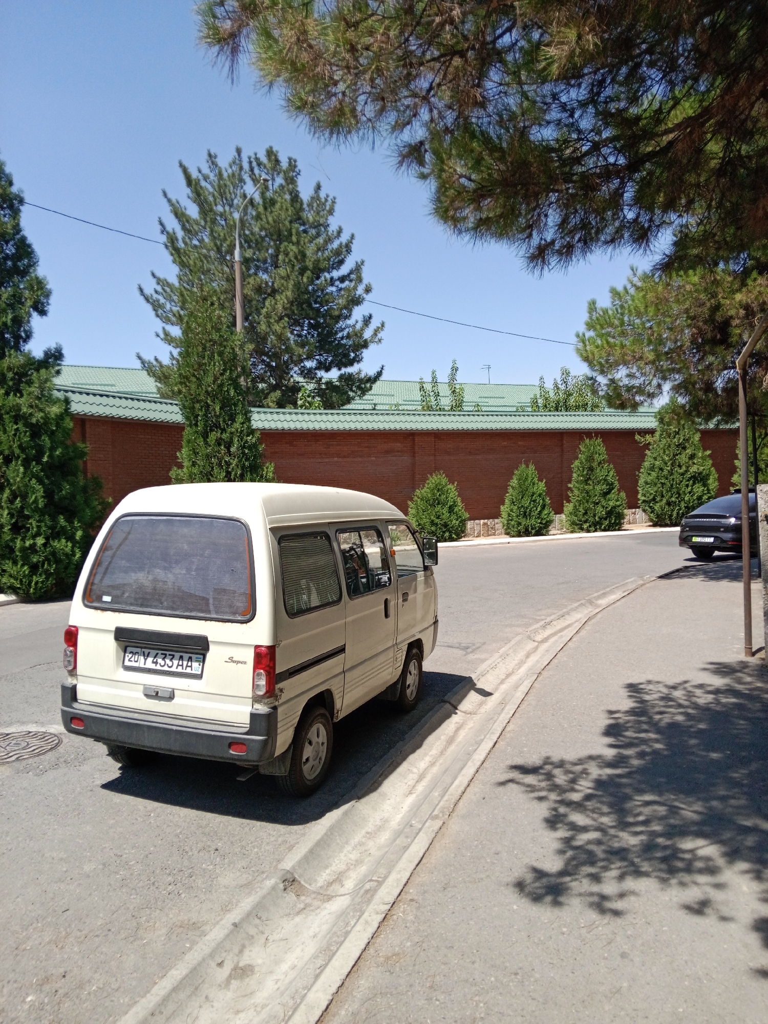 The image depicts a light-colored minivan parked on the side of a paved road in Tashkent, Uzbekistan. The minivan has a license plate that reads "20 Y 433 AA". Behind the minivan, a brick wall with a green-tiled roof runs along the property line. Several small evergreen trees are planted in front of the wall. The road curves to the right, and a dark-colored SUV is visible further down the road. Overhead, the branches of a pine tree cast shadows on the road. The sky is clear and blue. The scene appears to be an exterior shot, likely taken during the daytime.