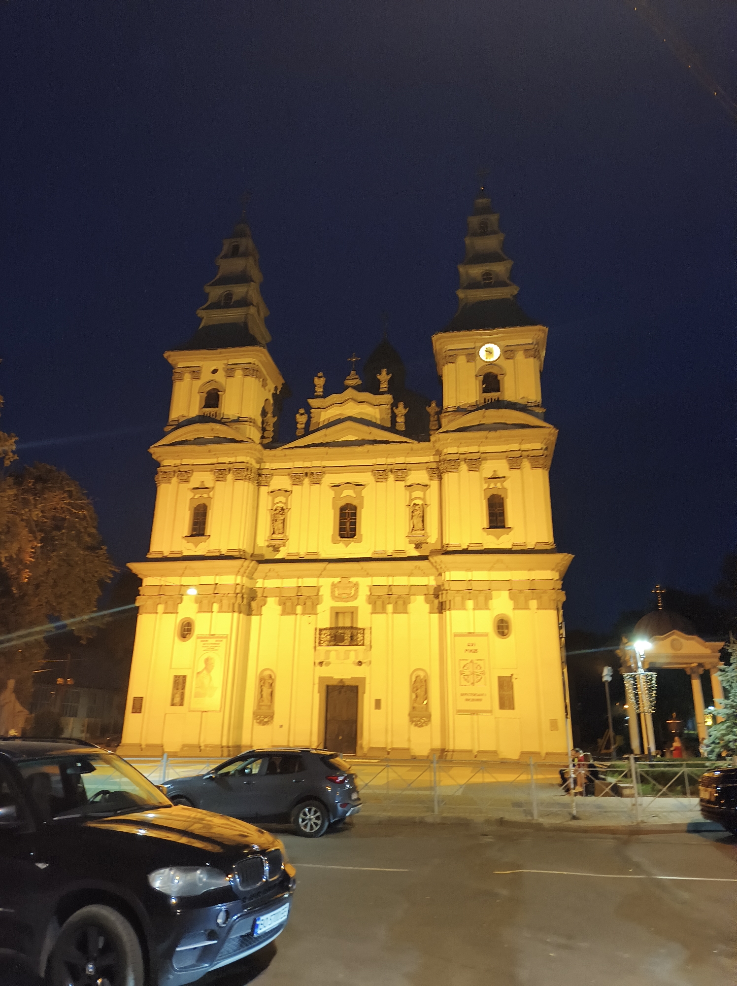 A night photograph captures the imposing facade of the Ternopil Dominican Church in Ternopil, Ukraine. The church, illuminated by artificial lights, displays a bright yellow exterior against a dark blue night sky. Its architecture features two prominent bell towers, each with multiple levels and ornate detailing, topped by crosses. The central section of the church has a large clock face on one of the towers and a series of windows and decorative elements. Below the main structure, there is a street with several parked cars. In the foreground, a dark BMW SUV is parked. Further back, a grey crossover SUV is also visible. A metal fence separates the church from the street, and beyond the fence, a few figures can be seen sitting. To the right of the church, a rotunda with a cross on top is illuminated, and a Christmas tree decorated with lights stands nearby. The scene suggests a public space, possibly a square or a street in front of a significant landmark, during nighttime hours.