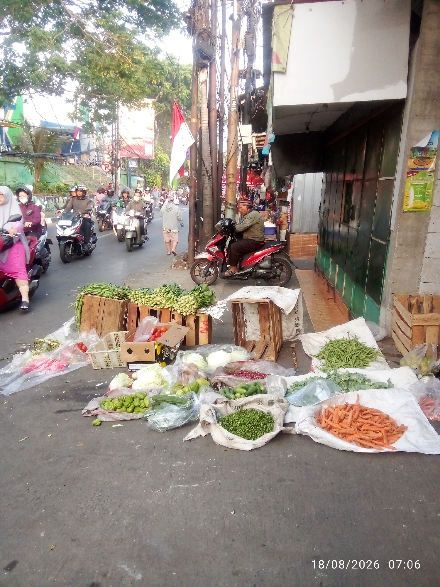 The image depicts a street scene in East Jakarta, Indonesia, with a focus on a makeshift market displaying various fresh produce. In the foreground, an assortment of vegetables is laid out on the ground and in wooden crates. These include carrots, green beans, peas, tomatoes, cabbage, and cucumbers, some packaged in plastic bags and others loose.
Several individuals are present, primarily on motorbikes, navigating the street. Some riders are wearing helmets and casual clothing, suggesting everyday commuting or market activity. A man in a light brown jacket and a patterned cap sits on a red motorbike in the middle ground, facing left. Other people on motorbikes are visible further down the street, moving in the same direction.
The environment appears to be an urban street with a concrete surface. Overhead, a tangle of electrical wires and poles dominates the upper portion of the image, characteristic of many Indonesian urban settings. A building with a green door and a white facade is visible to the right. A red and white flag, possibly the Indonesian flag, is mounted on a pole near the center of the image. Trees with green foliage line the left side of the street, partially obscuring some of the background structures. The lighting suggests it is daytime, possibly morning, given the timestamp "18/08/2026 07:06" overlaid in the bottom right corner.