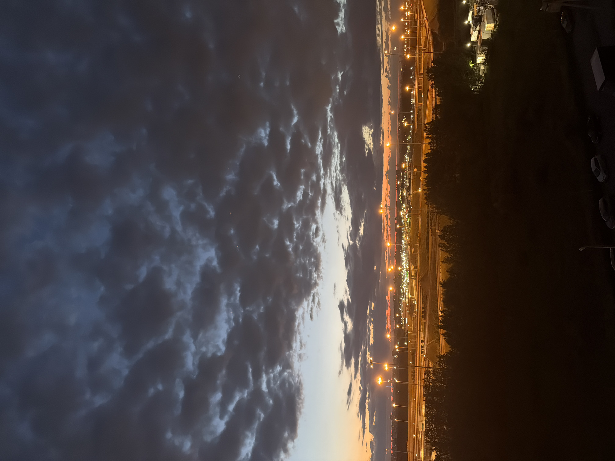 The image depicts a wide, horizontal view of an urban landscape at dusk. The sky is filled with dark, brooding clouds that are tinged with orange and blue from the setting sun near the horizon. A series of tall lampposts, illuminated with bright orange light, line a highway or roadway in the mid-ground. Beyond the roadway, a cityscape with numerous lights is visible, suggesting a developed urban area. A dark, silhouetted line of trees separates the foreground from the mid-ground. The foreground itself is largely obscured in shadow, but hints of parked vehicles, including cars and trucks, can be seen. The overall impression is of a transition from daylight to night in a busy urban environment.
