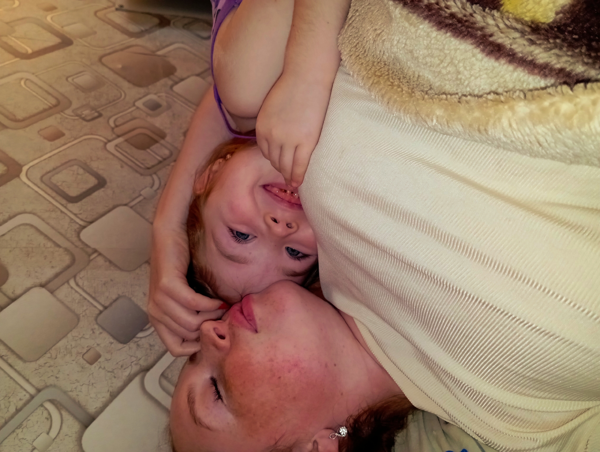 Two people, a woman and a child, are lying on a surface with patterned tile. The woman has freckles and an earring, and her eyes are closed. The child, with light-colored eyes, is smiling and looking up. A blanket is partially covering them. The location is Yurgamysh, Russia.