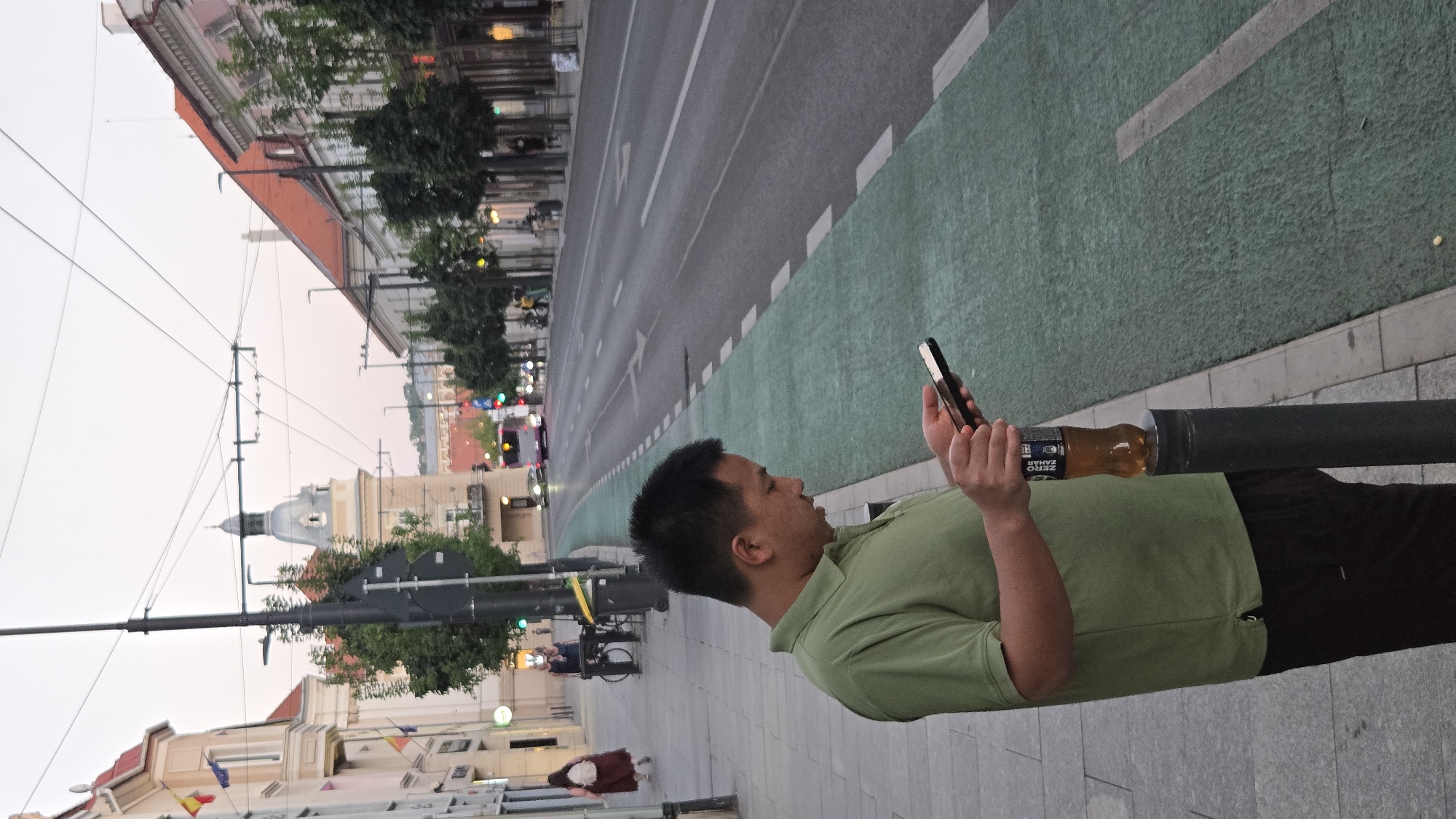 A man in a light green polo shirt and dark pants is standing on a sidewalk, looking at his phone. A bottle of "Zero Zahar" (zero sugar) soda is resting on a bollard next to him. The sidewalk has a green painted bike lane adjacent to it. In the background, there is a street with traffic, including a bus. Buildings line the street, and there are trees. The architecture suggests a European city. Based on the "Zero Zahar" branding, which is common in Romania, and the architectural style, the location is likely Cluj-Napoca, Romania, possibly on a street with public transportation lines visible overhead. The sky is overcast, indicating it might be early morning or late evening.