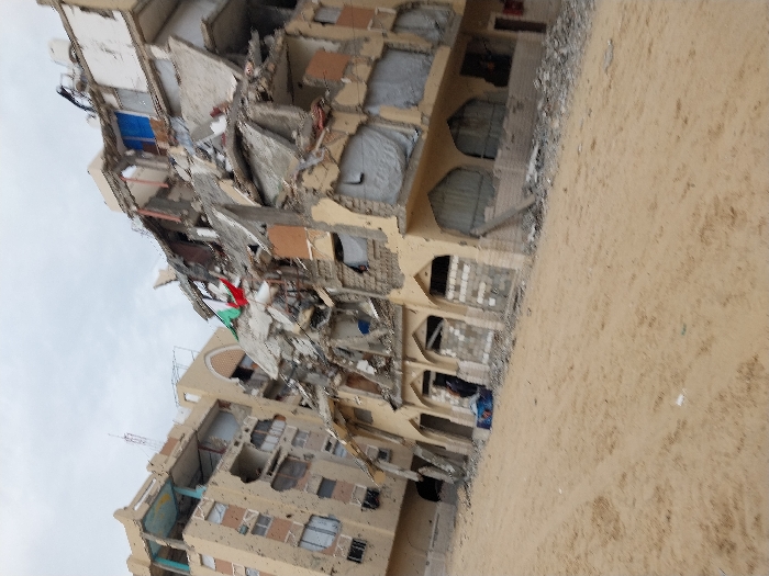 A severely damaged building stands under a cloudy sky. The structure is partially collapsed, revealing exposed interiors and debris. A Palestinian flag is visible amid the wreckage. The building appears to be multi-story, with several floors affected. The surrounding area is covered in debris and dirt, suggesting the aftermath of a bombing or other destructive event.