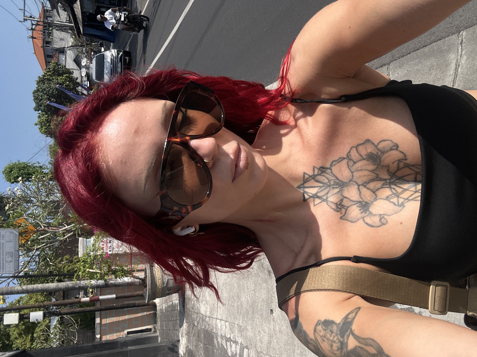 A woman is standing on a paved road in North Kuta, Indonesia. She has red hair, sunglasses, and a black top, and she has multiple tattoos on her body. She has wireless earbuds in her ears. There is a building to the left of the frame, and a street with a motorcycle and car in the background. The time of day appears to be midday with a clear blue sky.