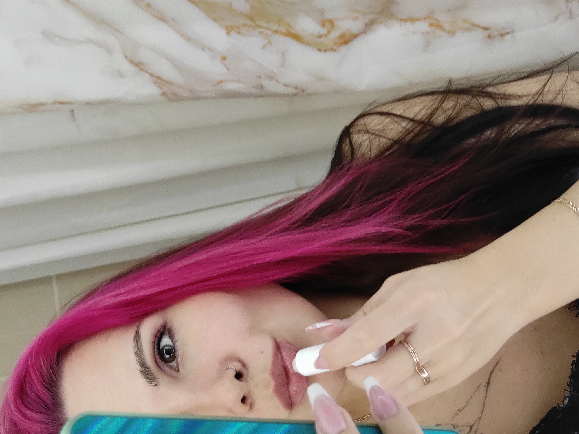 In this close-up, rotated image, a person with vibrant pink and dark hair is applying lip balm. Their face is partially visible, with one eye, nose, and mouth in focus. The person has a nose ring and is wearing makeup, including lipstick. Their hands, adorned with long, French-manicured nails and a ring on one finger, are holding a white tube of lip balm and applying it to their lips.
The background is a mix of textures. The upper portion shows a marbled surface with cream and gold tones, possibly a countertop or wall. Below this, there is a ridged, white surface, which could be part of the same structure or a different element in the setting. The lighting appears to be indoor and diffused, with no specific indications of the time of day or weather. The overall mood is intimate and personal, focusing on a moment of self-care or routine. The image is shot from a low angle, looking upwards towards the subject. There is no visible text in the image.