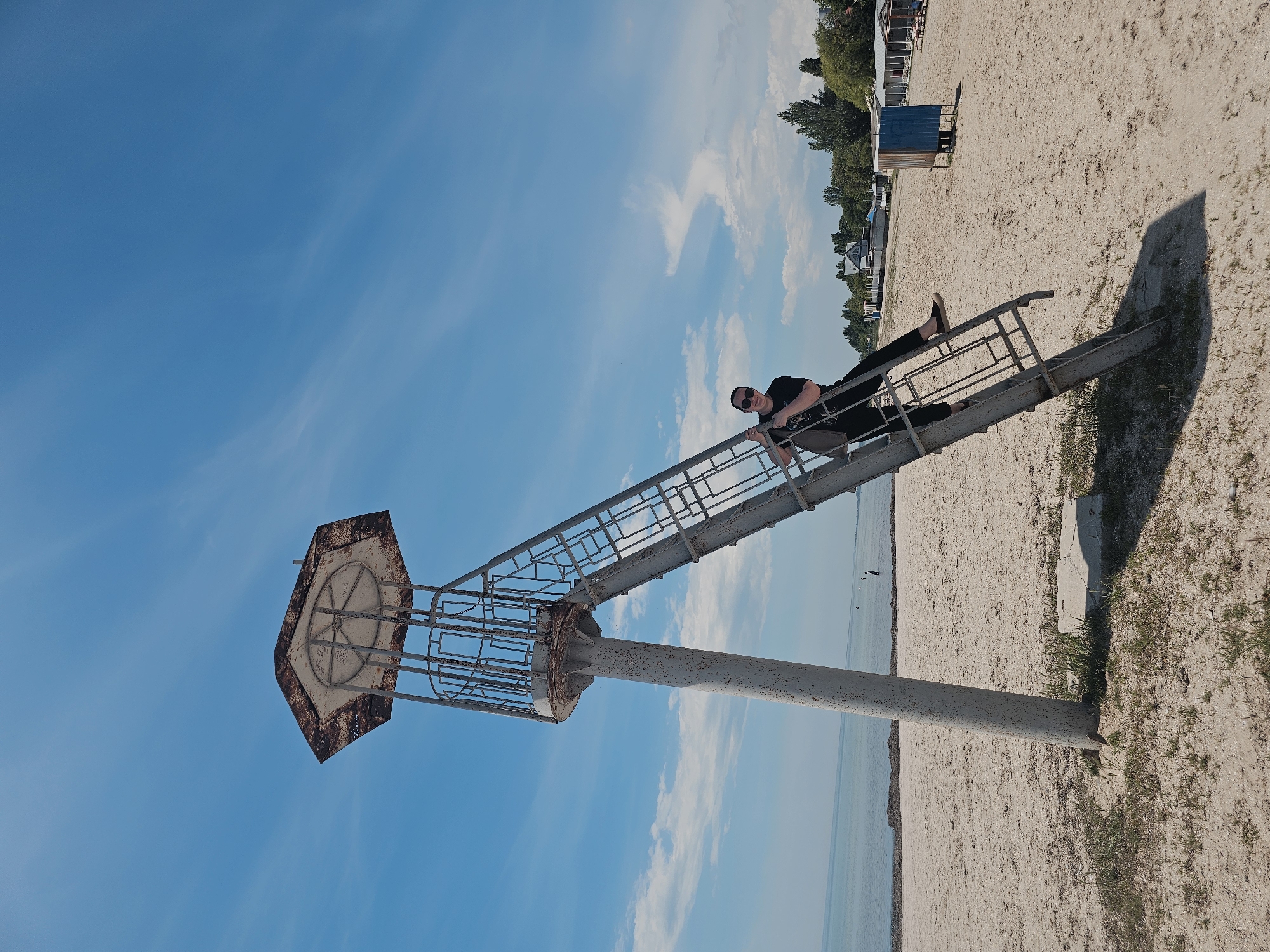 The image depicts a sandy beach scene in Skadovsk, Ukraine, under a clear blue sky with sparse white cirrus clouds. A weathered, light-gray metal lookout tower with an octagonal, rusted brown roof stands centrally, its main column anchored in the sand. A matching metal ladder, showing signs of rust and wear, extends from the sand upwards, leaning against the tower's column.
A person, wearing a black t-shirt, black pants, and sunglasses, is positioned on the ladder approximately halfway up. They are holding onto the ladder's rungs, facing slightly towards the camera with a subtle smile. Their left leg is bent, resting on a rung, while their right leg extends slightly downwards.
The foreground consists of light-colored sand with patches of sparse green grass and small weeds. The calm, light blue-green sea extends across the mid-ground, meeting the sandy shoreline. A visible line of dark seaweed runs along the water's edge. Several indistinct figures are observable in the shallow water in the distance.
To the right, further down the beach, a small, weathered blue and brown wooden changing cabin is present. Beyond it, a line of white-roofed structures, possibly additional changing rooms or beachside facilities, are partially visible, backed by a dense line of green deciduous trees and faint outlines of buildings, indicating the adjacent urban or suburban area of Skadovsk. The sunlight casts distinct shadows from the tower and ladder onto the sand.