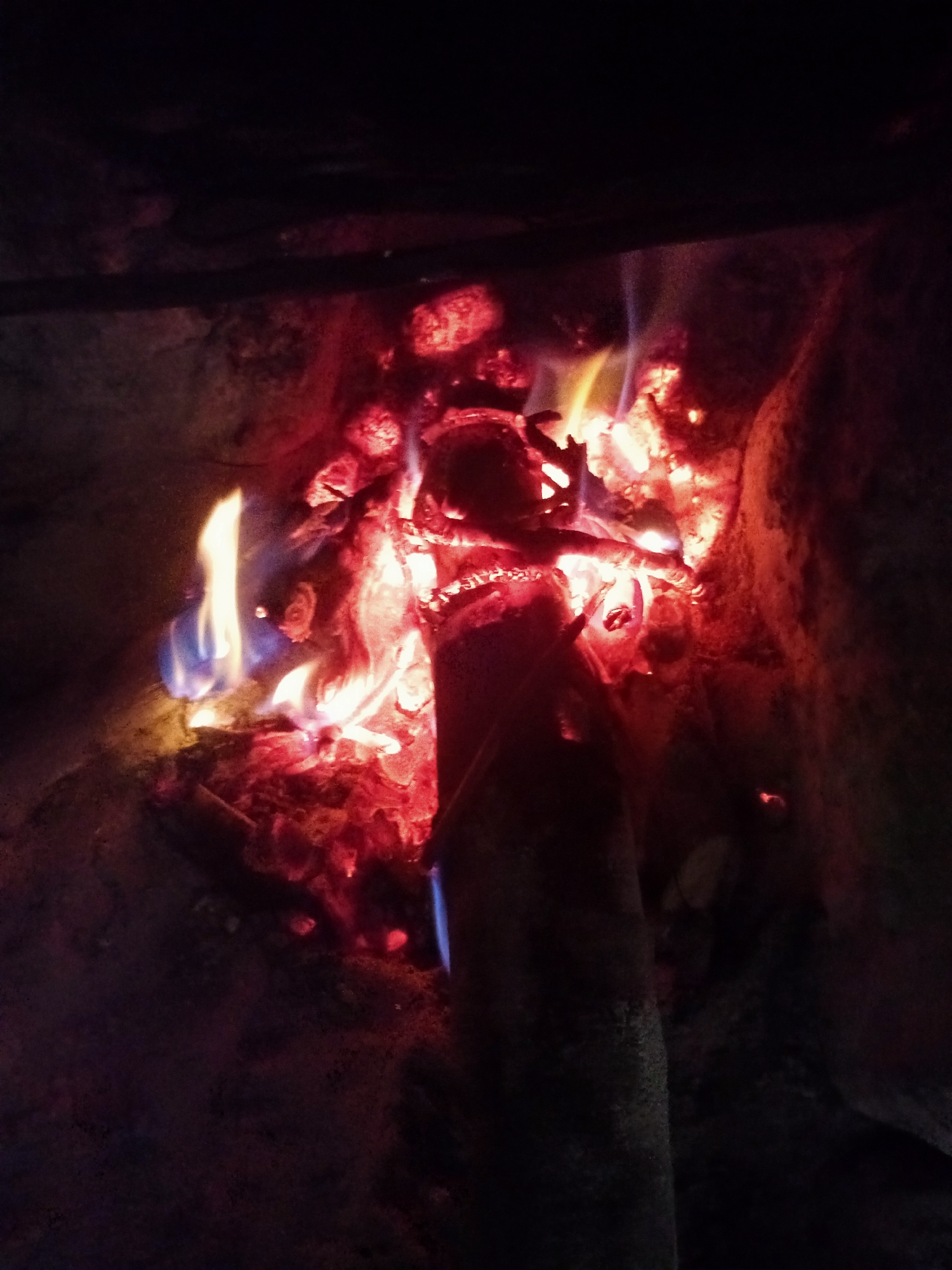 The image displays a close-up view of a burning fire, primarily showcasing glowing embers and flickering flames. The main subject is a rustic fire, with bright red and orange coals forming the base, while yellow and hints of blue flames emanate upwards from burning wood. A single, dark log is visible, partially consumed by the fire, extending from the bottom center into the fiery core.
The fire appears to be contained within a dark, textured enclosure, possibly a stone fireplace or a makeshift fire pit, suggested by the irregular, dark grey and brown surfaces surrounding the flames. The activity depicted is the natural process of wood combustion.
The setting is dark, indicating either nighttime or an interior space with minimal artificial lighting, making the fire the dominant light source. No people or visible text are present in the image. The scene evokes feelings of warmth, comfort, and primal energy. Considering the location context of Mugumo-ini ward, Kenya, such a fire might be used for cooking, warmth, or social gatherings. The colors are predominantly warm, with vivid reds, oranges, and yellows from the flames and embers, contrasted by the deep, dark tones of the surroundings.