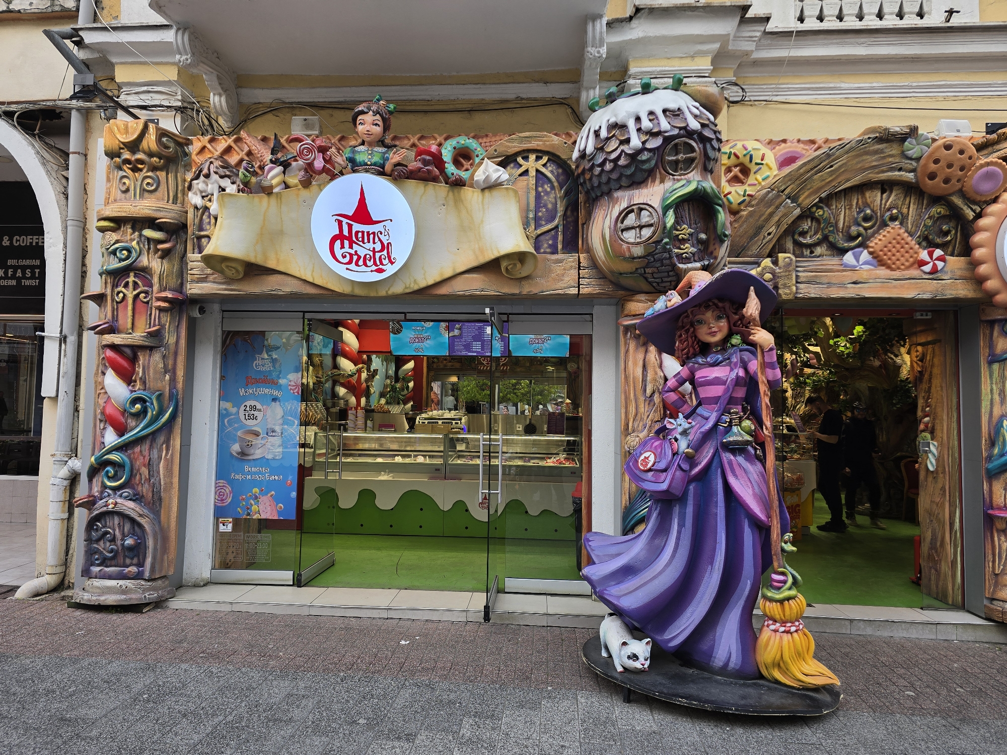 The image captures the storefront of "Hans & Gretel," a dessert shop located in an urban area. The facade is decorated in a whimsical, fairytale-inspired style with elements such as faux-wooden columns, confectionery decorations, and a house-shaped structure. A large figure of a witch, complete with a broom and a cat at her feet, stands to the right of the entrance. Through the glass doors, the interior of the store is visible, featuring display cases filled with various desserts. Customers can be seen inside the shop. Signage advertising special offers, such as discounts on specific products like "Доны" (likely "Donuts"), are posted on the storefront. A neighboring storefront, with a sign that reads "BREAKFAST," is partially visible on the left. The pavement outside the shop appears to be a mix of paving stones and asphalt.