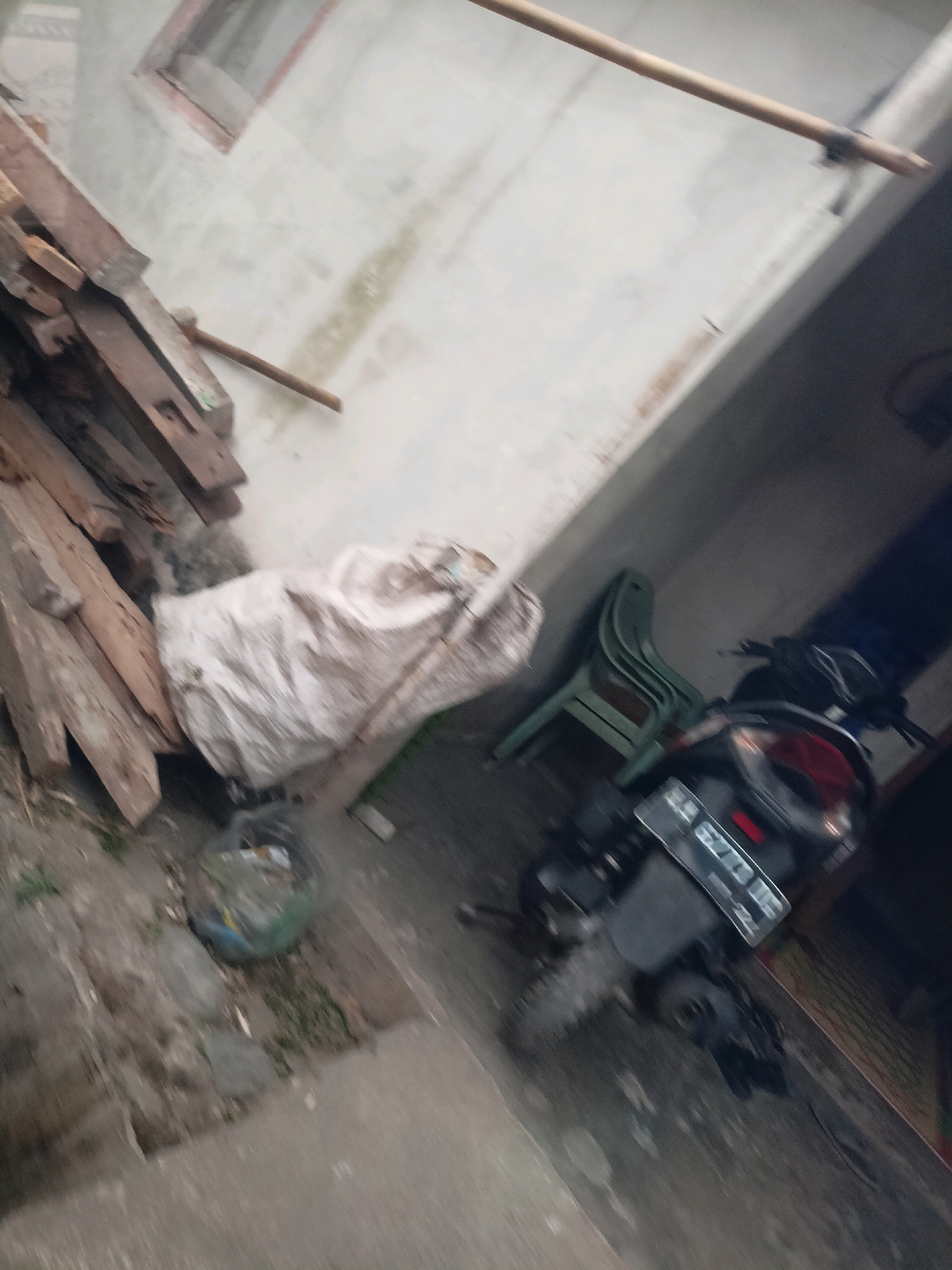 The image captures an outdoor scene in Kalegen, Indonesia, characterized by a slightly weathered white wall with a visible window frame. To the left, a pile of wooden planks is stacked, some appearing aged. Adjacent to the wood is a large, draped object that looks like a white sack or tarp, with a clear plastic bag containing debris beside it. In the mid-ground, a green plastic chair is leaning against the wall. A motorcycle is parked in the lower right portion of the image, with its license plate visible. The ground appears to be a mixture of concrete and dirt, with some sparse vegetation. The overall lighting suggests an overcast or dim environment, and the image quality is slightly blurred, making precise detail identification challenging.