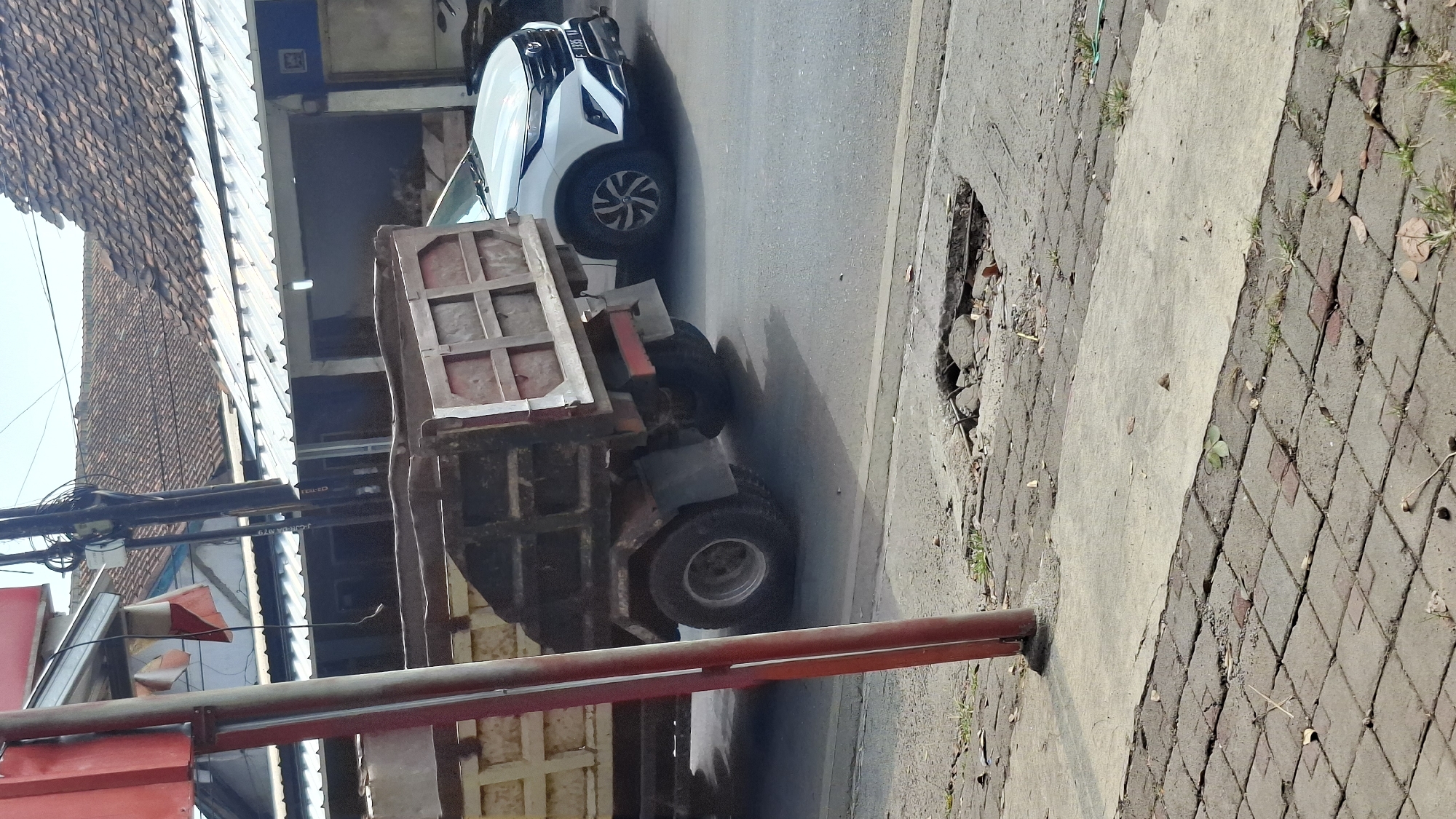 A medium shot captures a dusty street scene in Cilaku, Indonesia, under a bright, clear sky. In the foreground, a red pole stands to the left, and the paved sidewalk shows signs of disrepair with a large pothole in the road. A weathered dump truck, its bed filled with what appears to be sand or gravel, occupies the center of the frame, partially obscuring a white SUV parked on the right. The SUV, a Toyota Rush with license plate F 1335 VA, is positioned just behind the dump truck. In the background, traditional Indonesian houses with tiled roofs are visible, suggesting a semi-urban or suburban setting. The overall impression is of a typical day on a busy street, with a focus on the vehicles and the condition of the road.
