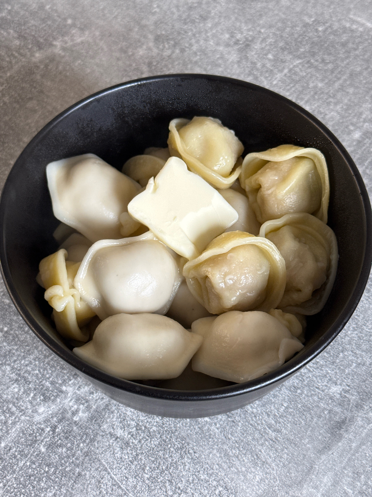 A black ceramic bowl filled with boiled dumplings, likely pelmeni or vareniki, sits on a textured gray surface, possibly a countertop or table. Several dumplings are visible, some with a dollop of white butter or sour cream on top. The dumplings appear to be a pale, translucent white with slight yellowing where they are cooked. The lighting is overhead, casting soft shadows. The environment suggests a home kitchen setting in Lyubertsy, Russia, based on the context provided. There are no people visible in the image.