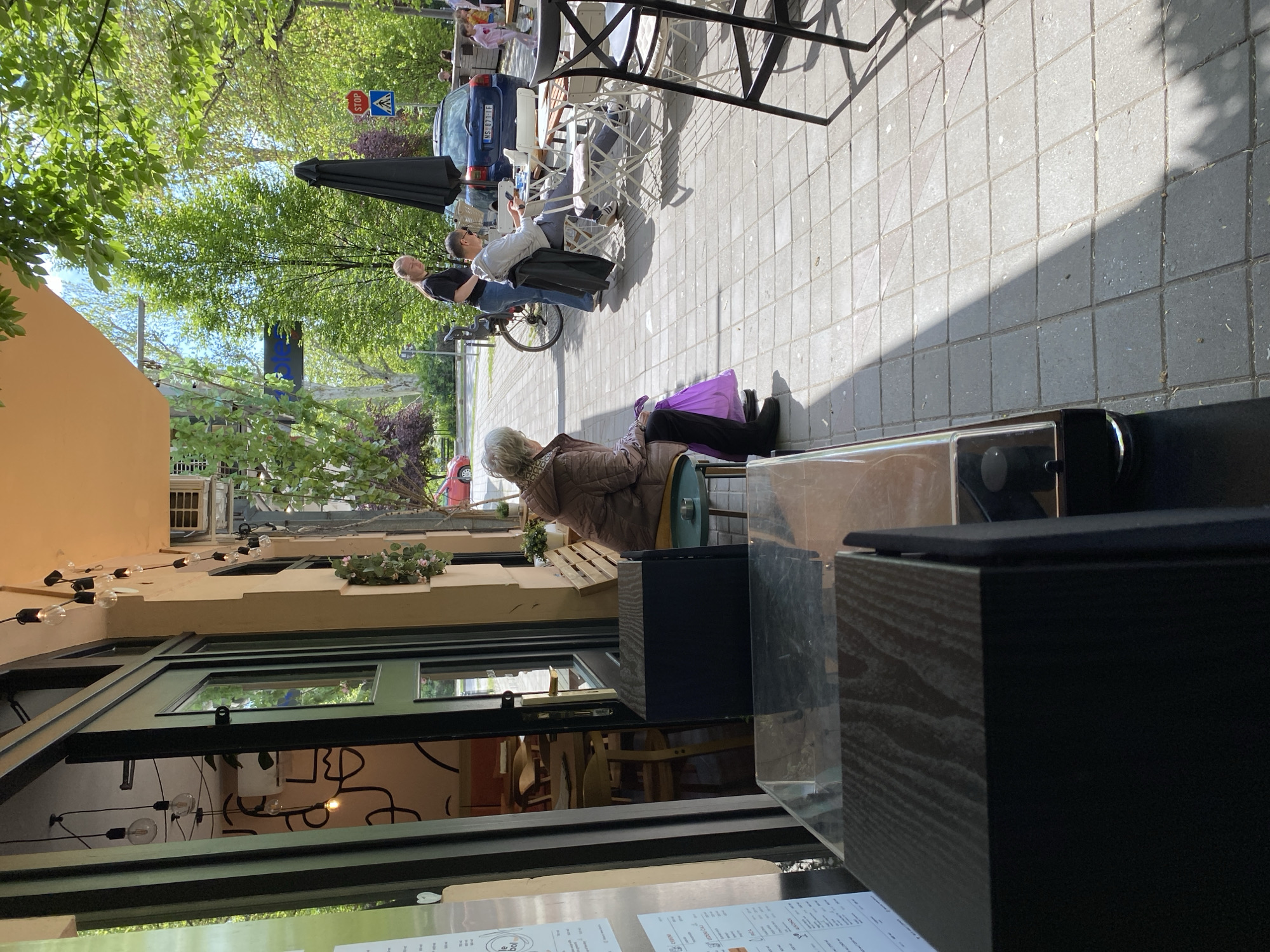 The image shows an outdoor cafe scene in Novi Sad, Serbia. Three individuals are visible on the sidewalk, with one person sitting at a table with an umbrella, while the other two are nearby. A parked car is visible in the background. A woman in a brown jacket and black pants is sitting on a green chair with a purple bag on the ground next to her. The foreground is occupied by the interior of the cafe, including a display case and menu, with tables and chairs visible through the window. Overhead string lights and decorative plants adorn the building facade.