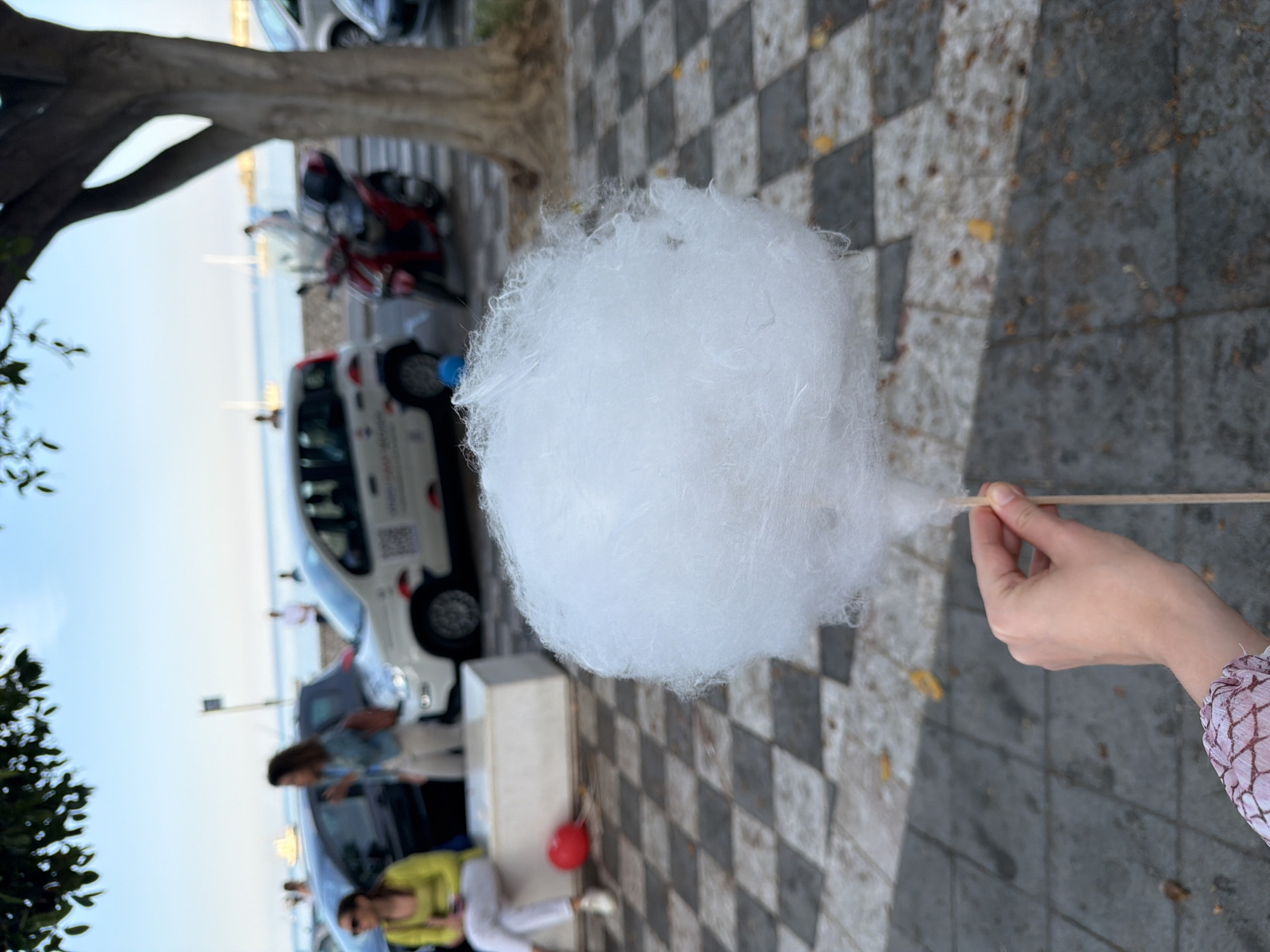 A close-up shot depicts a hand holding a large white ball of cotton candy on a stick. The person's arm is visible, clad in a pink and white patterned sleeve. The foreground features a black and white checkered sidewalk. In the blurred background, there are parked cars, people, and a tree. Based on the location and architectural details visible, the photo was taken on the lungomare of Chianchitta, Italy.