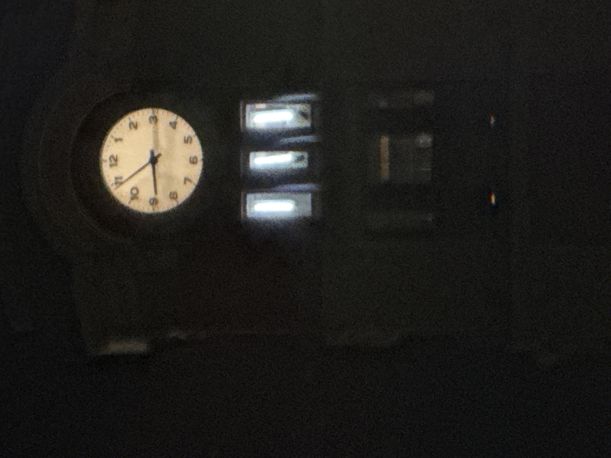 The image shows a dimly lit exterior scene at night. On the left, there is a round clock with a light-colored face and black numerals, showing the time around 8:18. To the right of the clock, there is a structure with three horizontal windows emitting a bright, bluish light. Further to the right, other structures are visible, with darker shapes. Two faint red lights are visible in the upper right. The overall environment is dark and grainy. The exact location in Varna, Bulgaria is not possible to ascertain without more information.