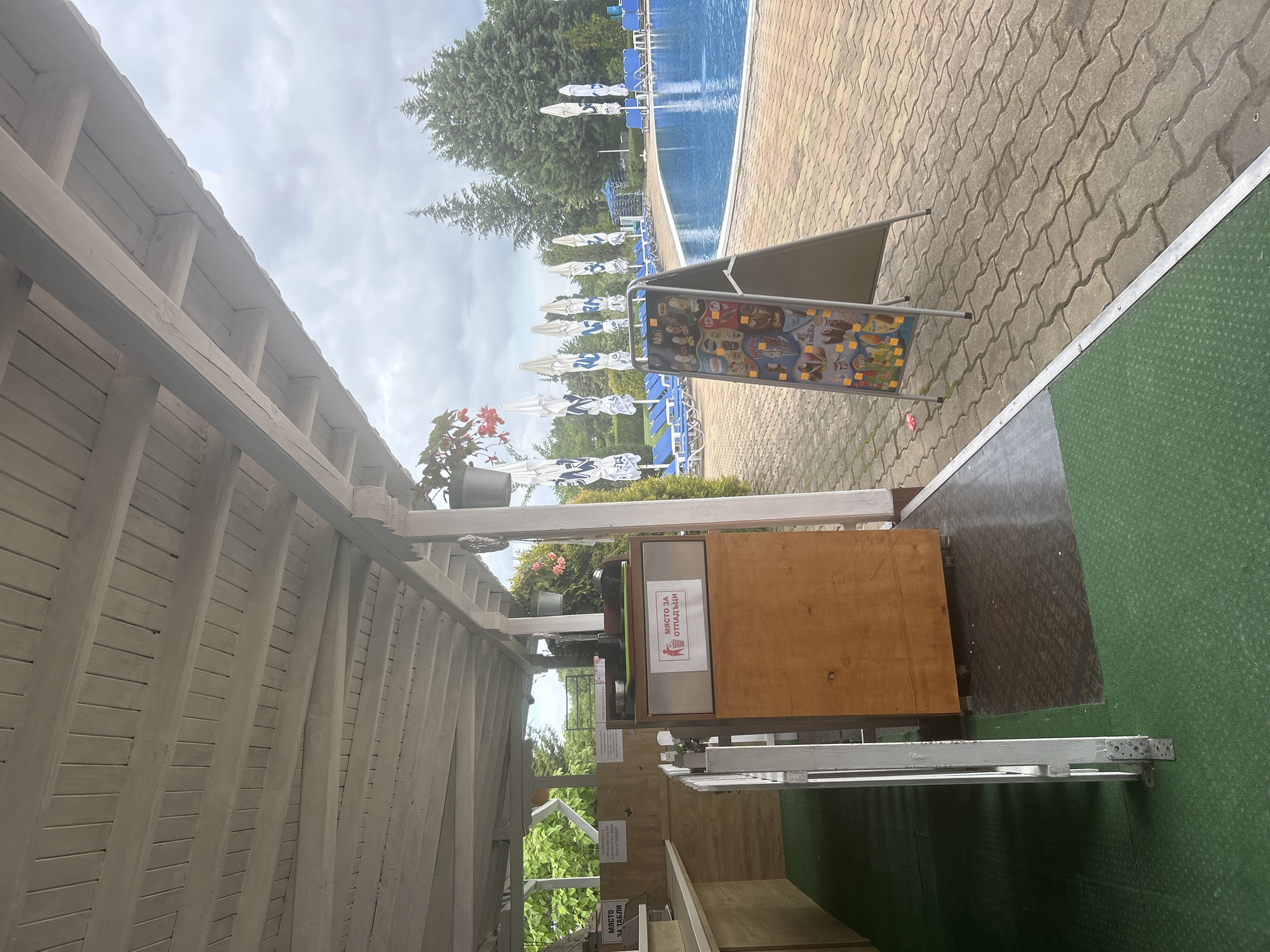 An outdoor scene at a swimming pool complex features a covered seating area with a white wooden ceiling and exposed beams. To the left, a counter area with wooden cabinets and a green mat on the floor is visible. A metal trash bin with a wooden facade and a sign indicating "МЯСТО ЗА ОТПАДЪЦИ" (Place for Waste) is to the right of the counter. Further to the right, a sandwich board displays an advertisement for ice cream. Beyond this, a blue swimming pool is surrounded by lounge chairs and large white umbrellas with blue logos. Evergreen trees form a backdrop against a cloudy sky.