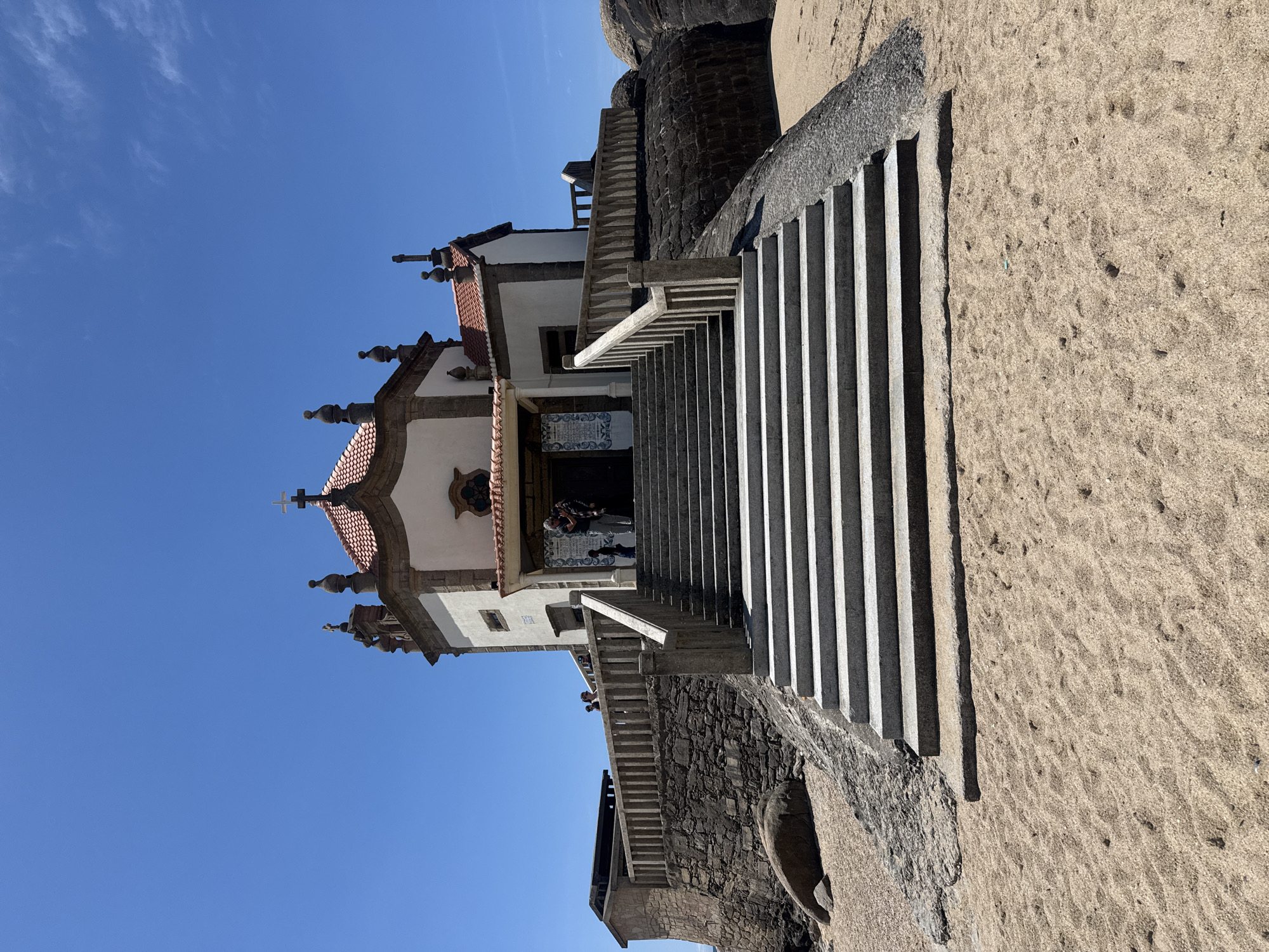 The image depicts the Chapel of the Lord of the Stone, also known as Capela do Senhor da Pedra, a small religious building located on Miramar Beach in Vila Nova de Gaia, Portugal. The chapel is characterized by its hexagonal shape and red-tiled roof, adorned with decorative pinnacles. A cross stands atop the chapel. The building is white with dark trim.
Several people are visible in the doorway of the chapel.
A long set of concrete stairs leads up to the chapel entrance. The steps cast strong shadows. To the right of the steps is sand. A dark stone structure is visible below and beside the chapel. The sky is a clear, bright blue. The image is taken at an angle, with the top of the chapel oriented to the left side of the frame.