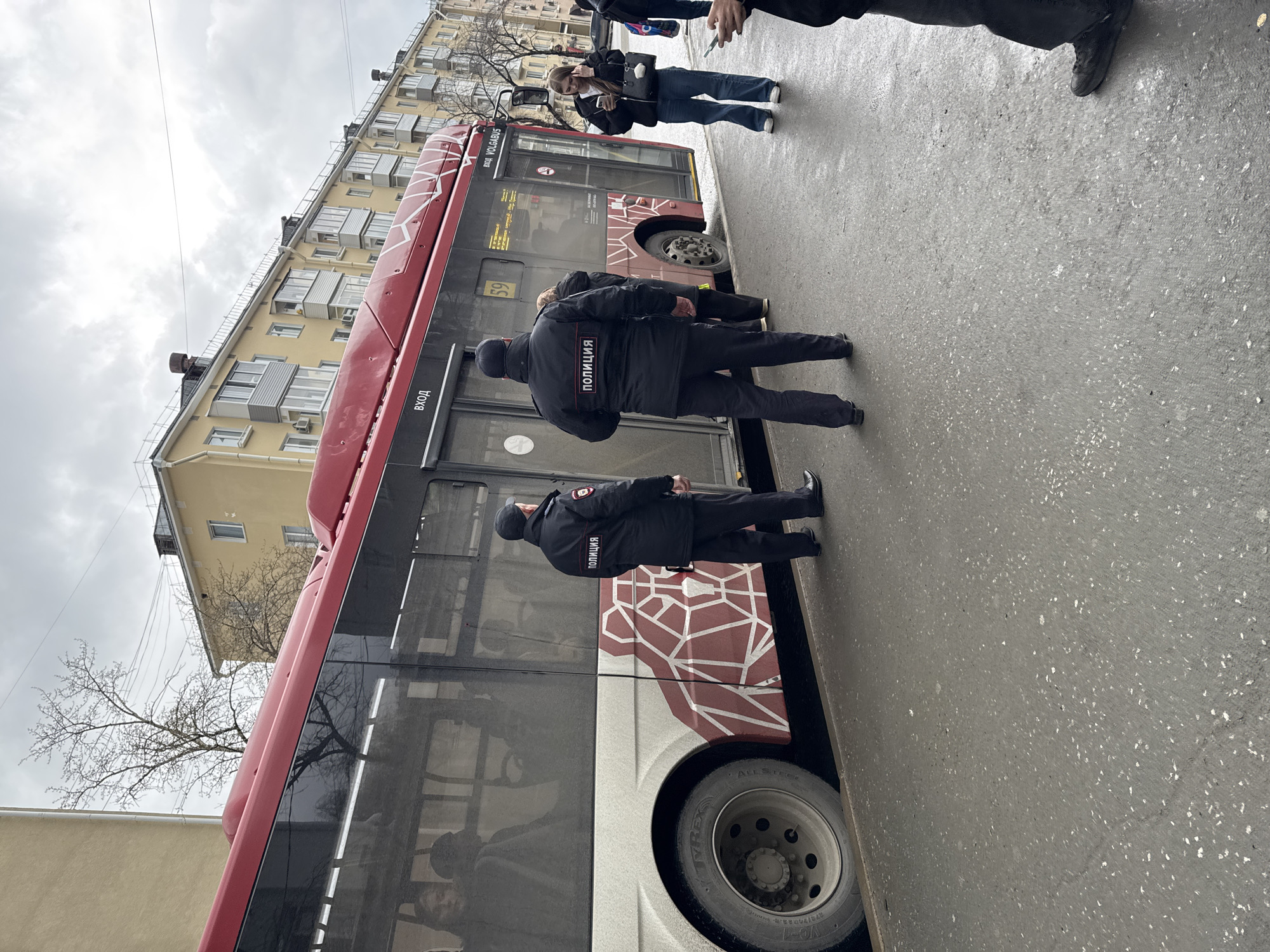 The image shows a city street scene in Perm, Russia. A maroon and grey Volvo bus is parked on the right side of the street near a yellow building. Two men in dark police uniforms with the word "ПОЛИЦИЯ" (POLICE) on their backs are standing near the bus door. Another person is near the door, partially obscured. Two people are standing across the street, one of whom is holding a phone and photographing the scene. The weather is overcast. The street is paved and appears to be slightly wet.