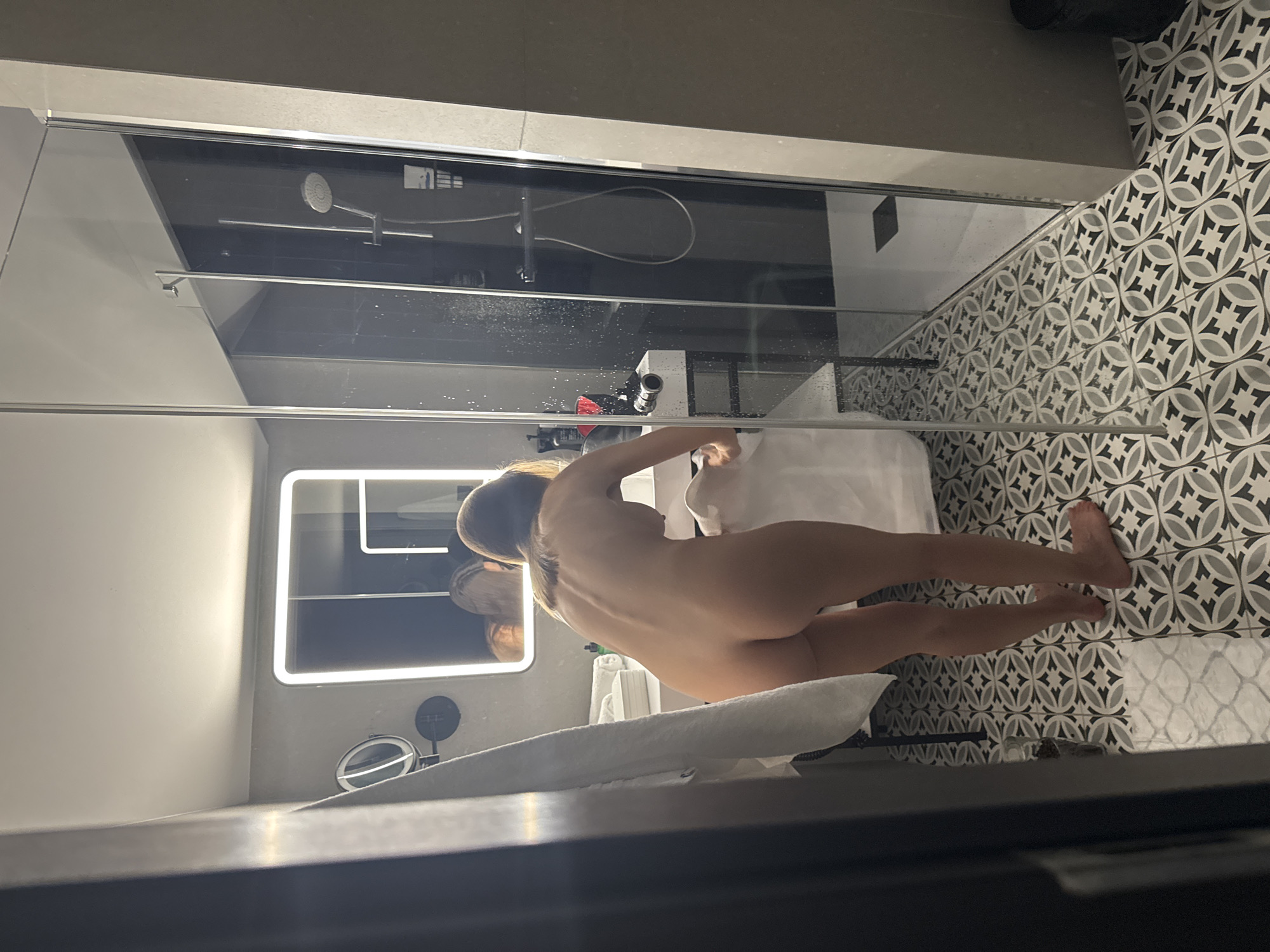 A medium-angle, vertically oriented photograph captures a person nude from the waist up, bending over a counter or sink in what appears to be a modern bathroom. The person is seen from the back, with their head slightly turned towards the right, revealing light brown hair pulled back. Their left arm is extended forward, reaching towards a white object on the counter, possibly a towel or basin. The posture suggests an action like washing or getting ready.
The bathroom features a patterned floor with grey, white, and black geometric tiles. To the left, a glass shower enclosure with a dark grey or black background is visible. Inside the shower, a modern showerhead and a horizontal bar are mounted on the wall. Water droplets are visible on the glass of the shower enclosure, suggesting recent use.
Behind the person, a illuminated rectangular mirror with a bright white light border is prominent on a light grey wall. A smaller, round mirror on a stand is also visible on the counter to the left of the person. A white towel is draped over the edge of the counter in the foreground, and other folded white towels are stacked behind the person on the counter. The overall lighting is artificial and bright, indicating an indoor setting, likely a hotel or a contemporary home. No discernible text is visible.
