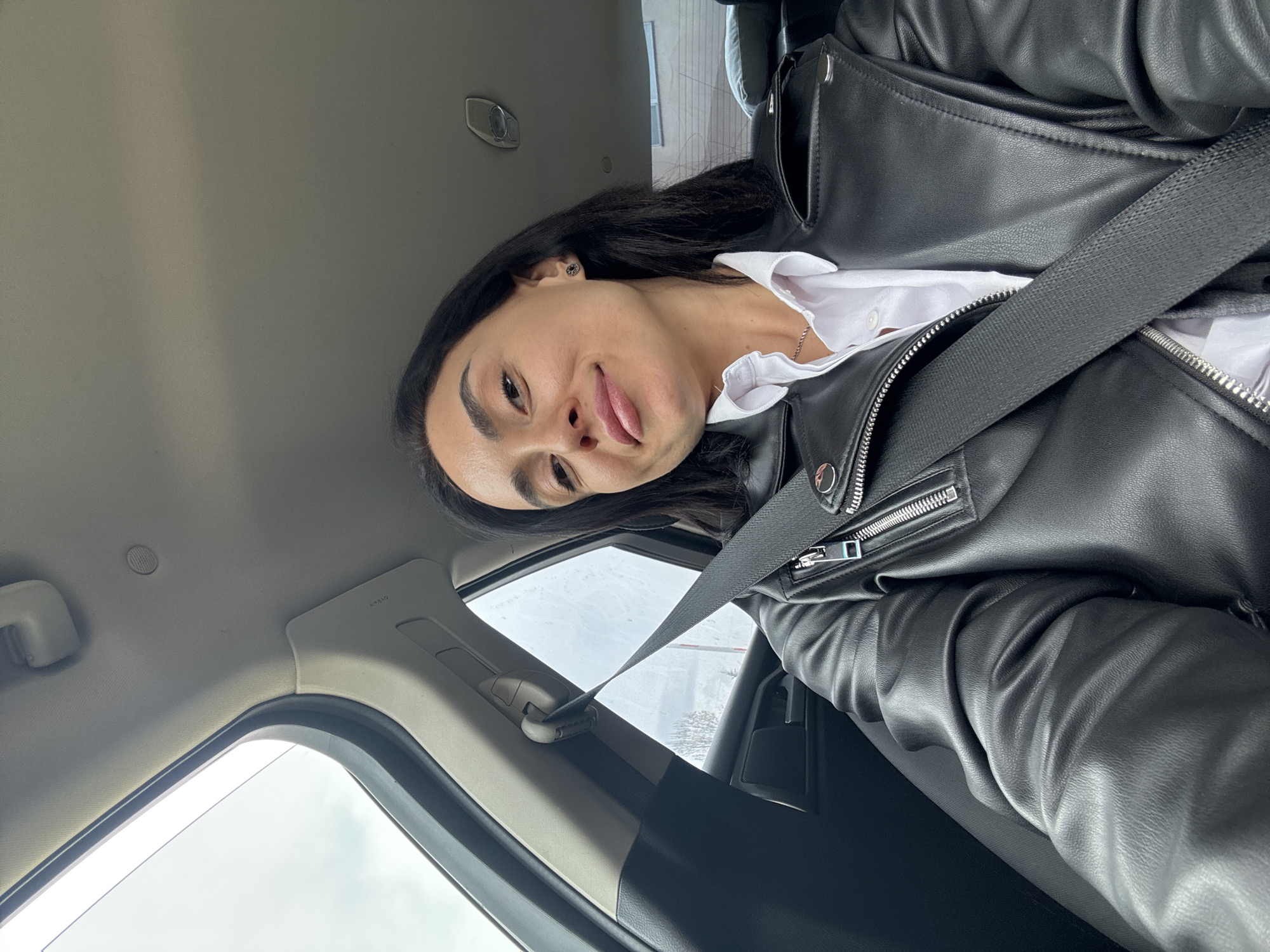 A woman is in a car wearing a black leather jacket and a white collared shirt. A seatbelt is fastened across her body. She has dark hair and is sticking her tongue out slightly. The background visible through the car window appears to be snowy or icy. The location is the city of Tbilisi, Georgia.