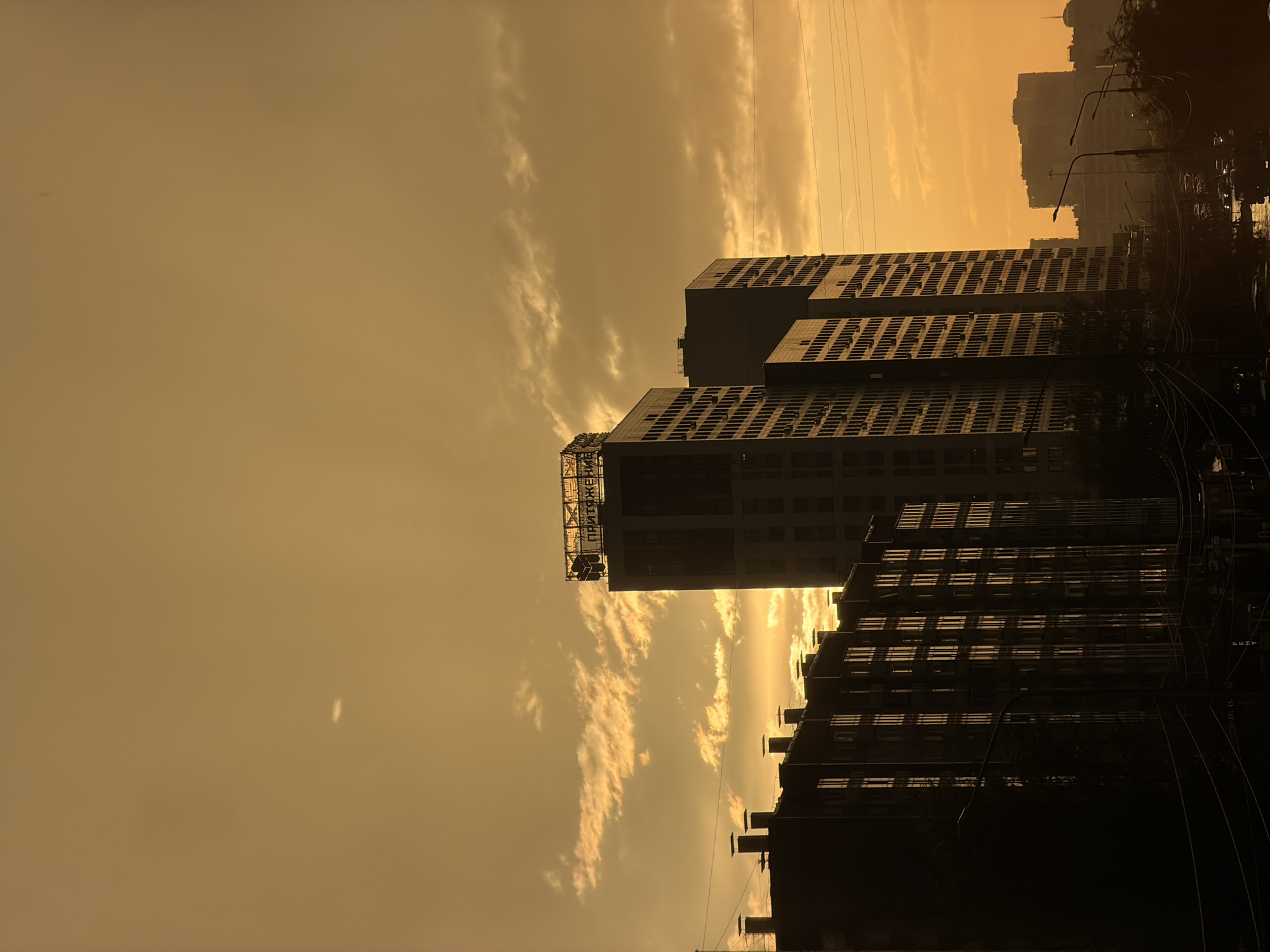 The image depicts an urban skyline at sunset in Krasnoyarsk, Russia. Tall, modern buildings with numerous windows dominate the scene, silhouetted against a warm, orange-tinted sky filled with scattered clouds. The foreground features a darker, more densely packed residential building with visible chimneys. Power lines crisscross the lower portion of the frame. In the distance, other buildings and streetlights are visible, suggesting a bustling city environment. The overall impression is one of a city at the end of the day, bathed in the soft, atmospheric light of a sunset.