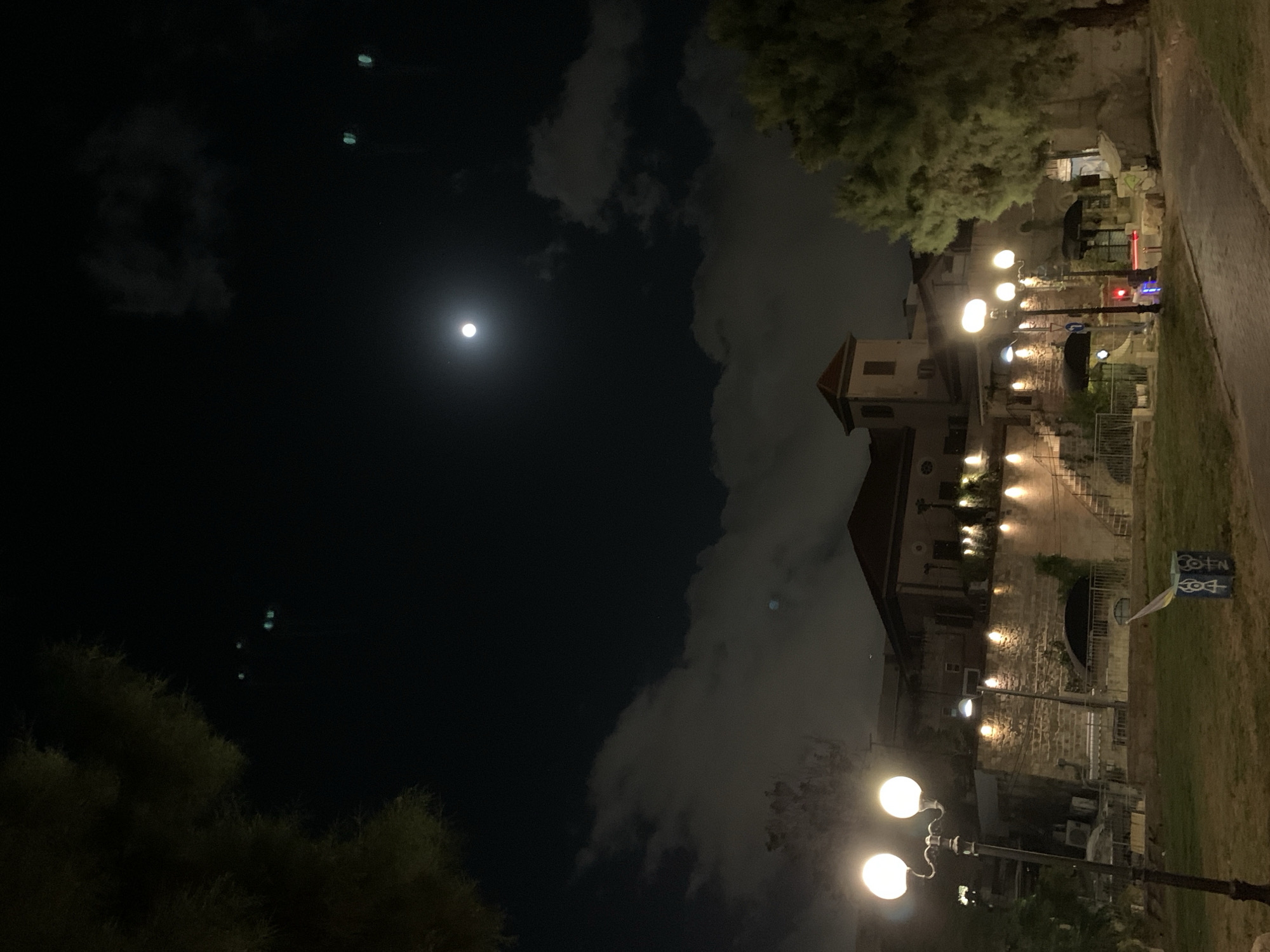 The image depicts a nighttime scene in Tel Aviv, Israel. A building, likely residential, stands on the right, illuminated by multiple street lamps. The structure has a light-colored facade and appears to be constructed of stone or brick. A tower-like feature is visible on the roofline. Vegetation, including trees and shrubs, surrounds the building and pathway. The sky is dark with scattered clouds and a bright full moon. A street sign is also visible. The scene is captured at night with artificial lighting and natural moonlight providing illumination.