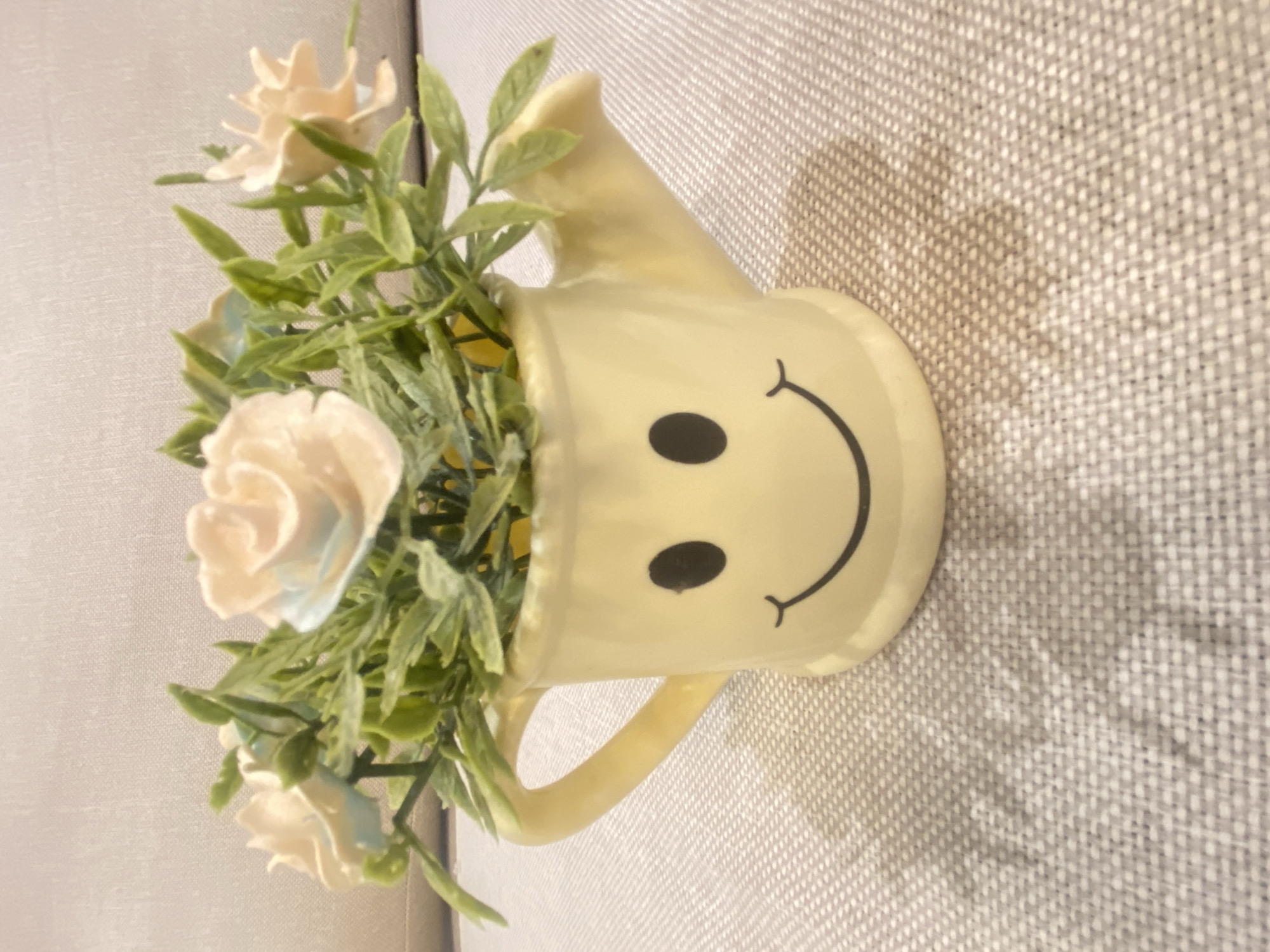 A slightly off-center, overhead shot displays a small, light yellow watering can-shaped container with a simple smiley face drawn on its front. The container holds artificial plants with small white roses and green leaves. The object sits on a textured, light gray surface. There is no evidence to indicate that the photo was taken in Alanya, Turkey. There are no people, subjects, or interactions visible.