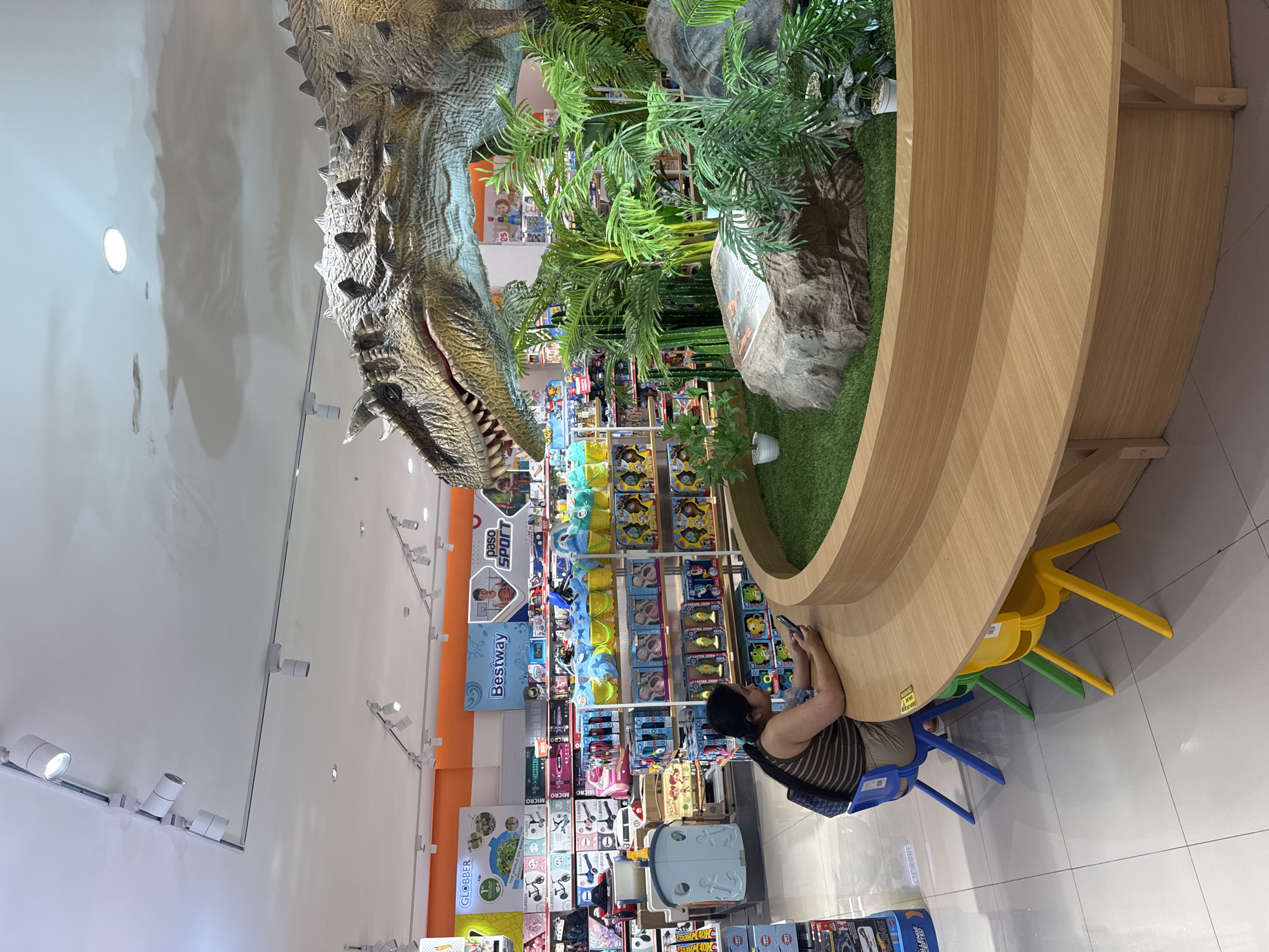A woman with dark hair, wearing a striped tank top and beige shorts, sits at a curved wooden bench in a toy store in Kuta, Indonesia. She is looking down at her smartphone, with her left arm resting on the bench. In front of her, several colorful children's chairs are visible: one blue, one green, and one yellow. Behind the bench, a large, realistic model of a dinosaur is displayed amongst artificial plants and rocks, with a grassy area in front of it. The background of the store shows shelves stocked with various toys and merchandise. The ceiling features a track lighting system.