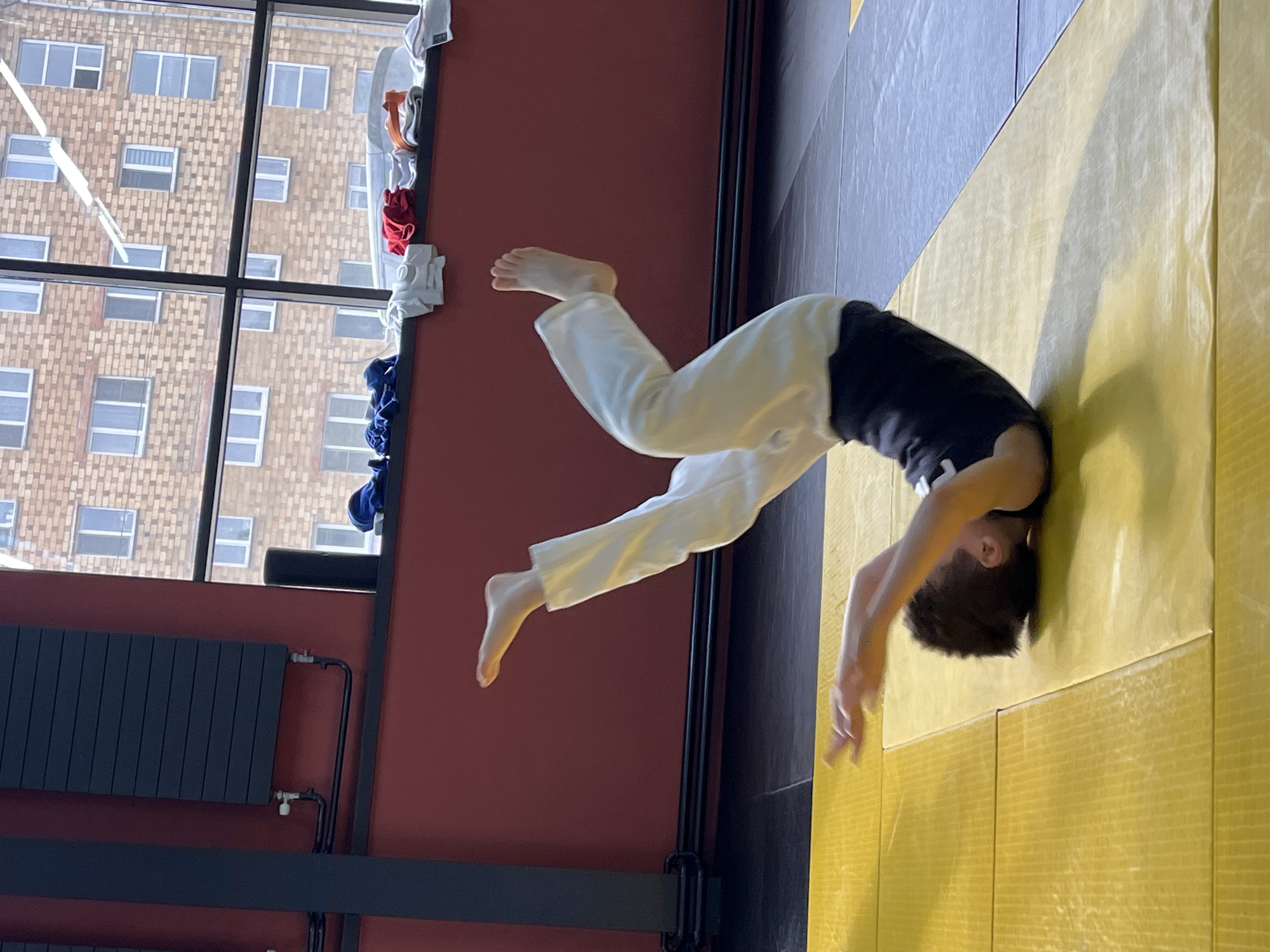 An indoor scene in Krasnoyarsk, Russia, shows a young person in mid-action, seemingly performing an acrobatic maneuver or martial arts move. The individual, dressed in a black t-shirt and white pants, is inverted with their head close to a yellow padded floor mat and their legs extended upwards in the air. Their bare feet are visible.
The setting appears to be a training facility or gym. The floor is covered with bright yellow padded mats. A prominent red wall runs along the left side of the frame, contrasting with a darker, possibly black or dark grey, section on the right. A large window occupies the upper left portion of the wall, revealing the exterior of a multi-story building constructed from light brown bricks, featuring numerous rectangular windows. Bright daylight streams through this window, indicating it is daytime.
Along the right edge of the window frame, various folded items of clothing or fabric in white, orange, red, and blue are neatly stored. In the lower left corner, a dark grey panel radiator with visible pipes is mounted on the red wall. Black horizontal and vertical beams are also visible, possibly part of the window structure or room architecture. No legible text is visible in the image. The scene captures a dynamic moment of physical activity.