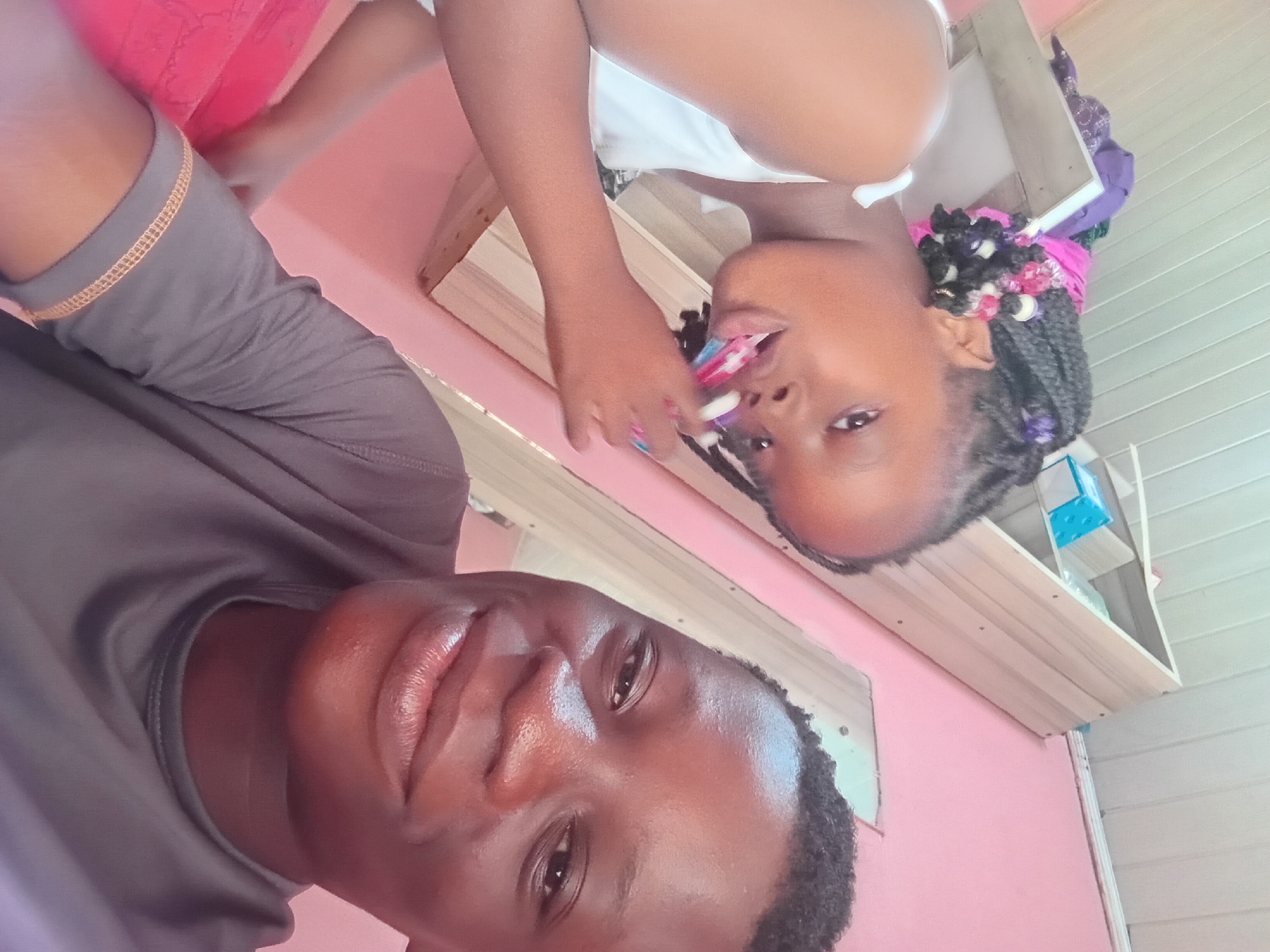 Two young Black individuals are posing for a selfie in an indoor setting. The person in the foreground on the right is an older youth with short, dark hair and is wearing a grey t-shirt. They are smiling directly at the camera. Behind them and to the left, a younger girl with braided hair is partially visible. She is wearing a white tank top and appears to be brushing her teeth with a colorful toothbrush. In the background, there are shelves with some items on them and a pink wall. The environment seems to be a domestic interior.