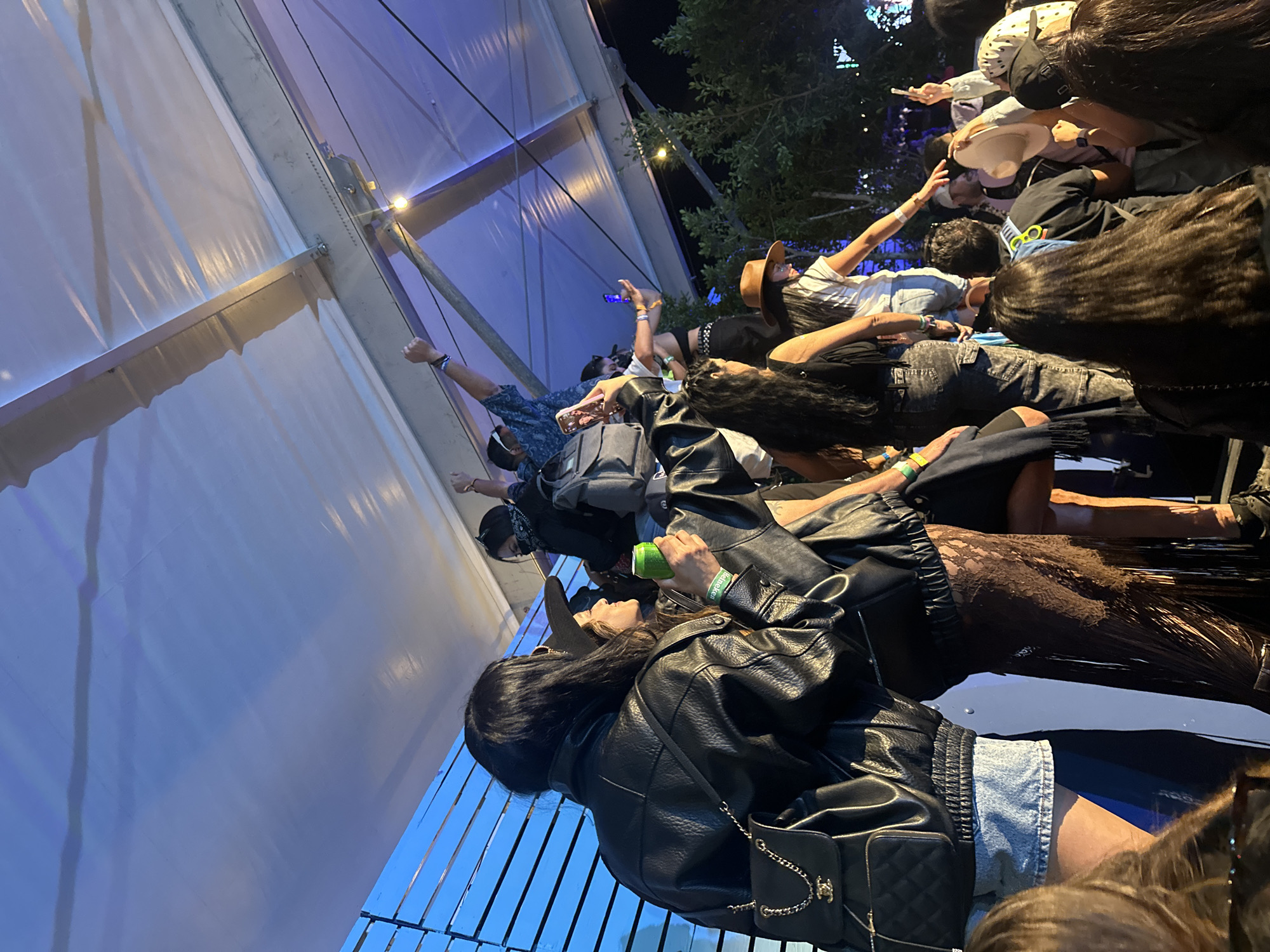 The image shows a crowd of people, likely at a concert or festival, with a large white tent structure visible in the background. People are standing close together, some with arms raised. One person in the foreground is wearing a black leather jacket and denim shorts, holding a green can. The location appears to be at night, with a mix of natural and artificial lighting. There are trees visible in the background.