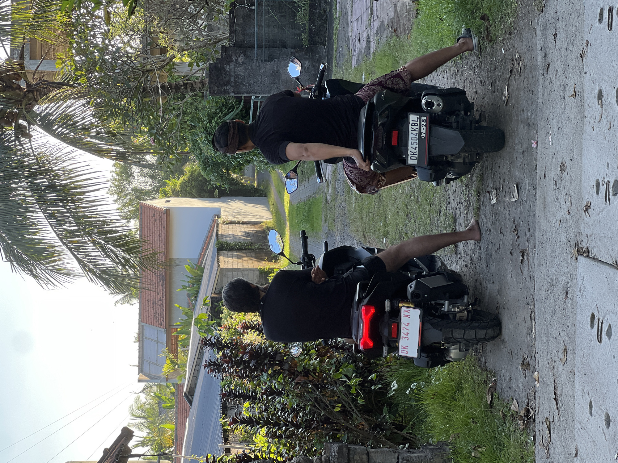 Two men are depicted from behind, sitting on black motorbikes that are parked on a paved or dirt path. The scene takes place in a sunny, tropical environment, likely in the Ubud District of Indonesia, given the context and visual cues.
The man in the foreground is closer to the camera, wearing a dark short-sleeved shirt and dark shorts/pants, with his feet bare. He is positioned on a black scooter with a red taillight. The license plate visible on his motorbike reads "DK 3474 XX".
Further down the path, the second man is also seated on a black Honda scooter. He wears a dark short-sleeved shirt, a dark headband, and patterned shorts. One of his feet is bare, while the other has a dark sandal on. The license plate on his motorbike displays "DK 4504 KBL" and "07-28" underneath. The "Honda" brand name is also visible on both bikes.
The surrounding area is lush with tropical vegetation, including large palm trees on the left and various bushes and plants along the path. In the background, simple buildings with tiled roofs can be seen under a bright, clear sky, suggesting either late morning or late afternoon with strong sunlight casting some shadows. The ground is a mix of concrete, dirt, and sparse grass. The overall atmosphere appears calm and warm.