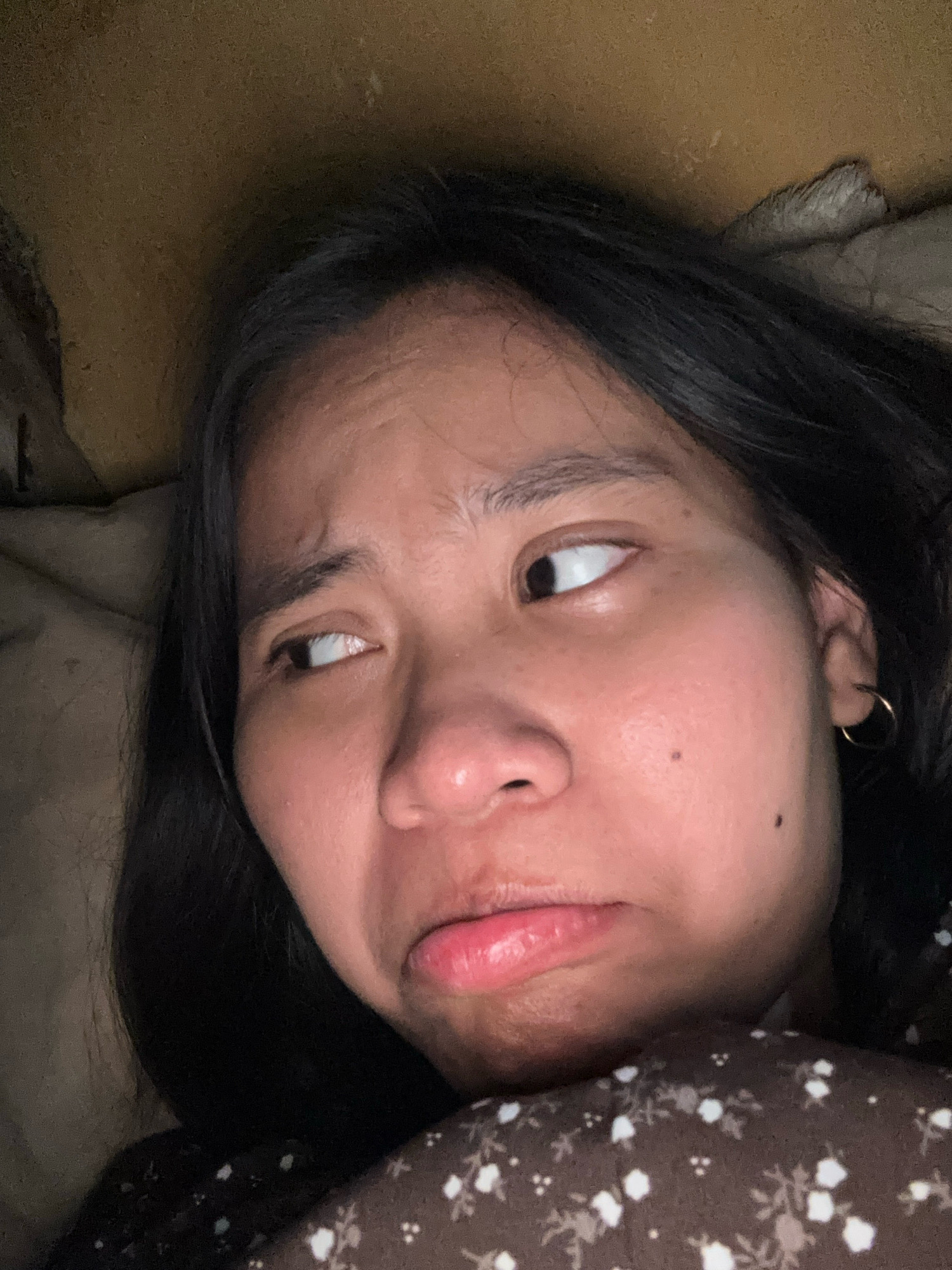 A close-up, low-angle, indoor selfie shows a young East Asian woman lying down, with her face angled to the left and looking away from the camera. Her dark hair frames her face. She has a neutral to slightly displeased expression, with her eyebrows furrowed and her mouth slightly pursed. A small, gold hoop earring is visible in her right ear. The background is a dark, textured surface, possibly a bed or pillow, with a faint floral pattern on a dark fabric visible at the bottom right of the frame. The lighting is dim, creating shadows on her face. The location is East Jakarta, Indonesia.