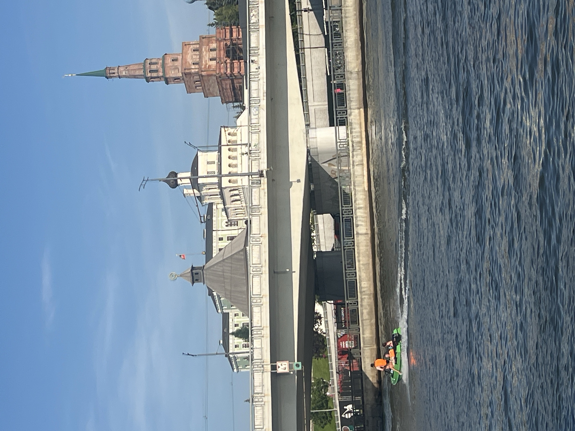 This image shows a view of the Kazanka River in Kazan, Russia, with the Söyembikä Tower and the Kul Sharif Mosque in the background. In the foreground, a small green inflatable boat is navigating the river, carrying at least three individuals, two of whom are wearing bright orange life vests. The boat is leaving a wake behind it, indicating movement. A large concrete bridge spans the river in the mid-ground, with a sign displaying the number "47" hanging from its underside. Behind the bridge, several historical buildings, including a domed structure and a tall, multi-tiered tower with a green spire, are visible against a clear blue sky. The water is rippling, reflecting the sunlight.