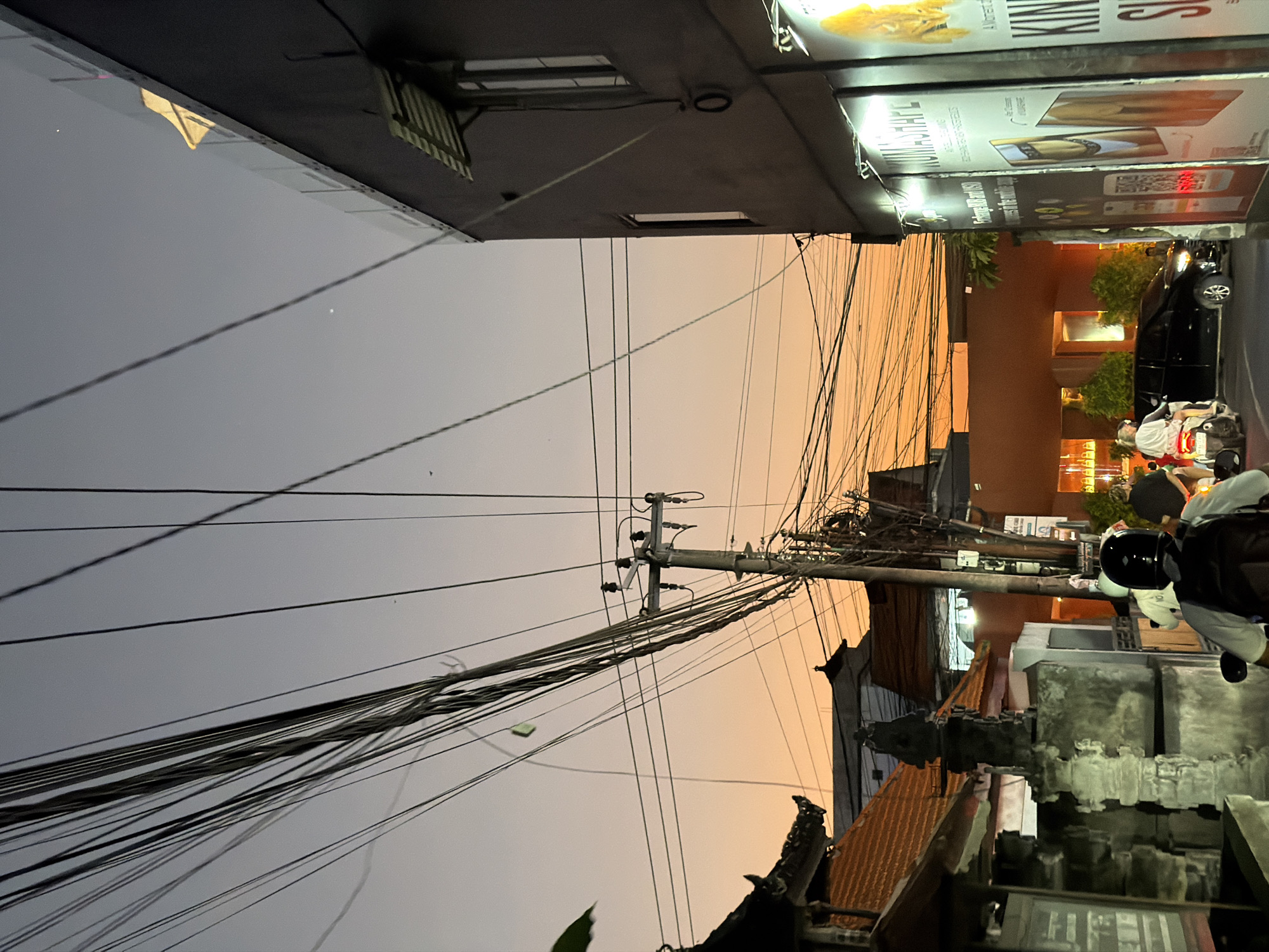 The image depicts a twilight street scene in North Kuta, Indonesia, characterized by a gradient sky and a dense network of overhead power lines. The sky transitions from a soft orange at the horizon to a muted grey-blue overhead. Numerous black power lines crisscross the upper and middle sections of the frame, originating from a utility pole positioned centrally.
On the left, traditional Balinese-style roofs with terracotta tiles are partially visible, alongside a grey stone pillar or shrine structure in the foreground. Green foliage is present on the far left.
On the right, a modern, dark grey multi-story building occupies the upper and middle sections. It features a glass-paneled balcony railing on its upper floor and vertical windows. Below, the building's facade displays several brightly illuminated vertical advertisements. One panel clearly shows "KUMASHAPE" with accompanying text and images, including a prominent cat illustration. Another visible panel mentions "Exchange HKD and USD" and "KING SIZE".
In the lower foreground, three individuals are riding two scooters away from the camera. The closest individual wears a black helmet and a white shirt with a black backpack. Directly behind them, another person on a scooter wears a white top and a white helmet. A black multi-purpose vehicle (MPV) is positioned slightly further down the street, its headlights visible. The street itself appears to be paved, with additional light sources emanating from building interiors and street fixtures.