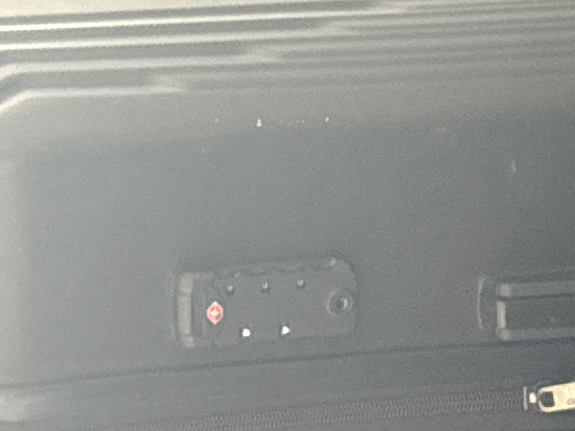 The image shows a close-up of a black suitcase. On the surface of the suitcase, there is a TSA-approved luggage lock with three rotating dials. The lock is rectangular and black, with a red diamond-shaped symbol on it. Below the lock, there are visible seams and stitching details of the suitcase. The lighting suggests an indoor or shaded environment, and the background is blurred, indicating a shallow depth of field. The overall scene is focused on the detail of the luggage lock.