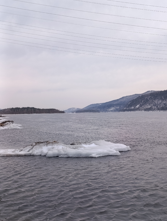 This image depicts a vast, cold-weather riverine or lake landscape located in the city of Слизнево, Russia. The foreground and midground are dominated by a wide expanse of dark grey, rippling water. Centered in the midground is a large, irregularly shaped ice floe, predominantly white with some visible darker, possibly rocky, surfaces. Smaller accumulations of white ice are present along the distant left bank. The background features a continuous body of water extending towards the horizon. To the far left, a low-lying, dark-forested landmass or island is visible. To the right, a range of snow-covered mountains or large hills rises, displaying numerous dark coniferous trees on its slopes. The sky above is uniformly overcast and light grey. Multiple parallel power lines traverse the upper third of the frame horizontally. No individuals, animals, or active subjects are visible within the scene. The environment indicates a late winter or early spring setting.